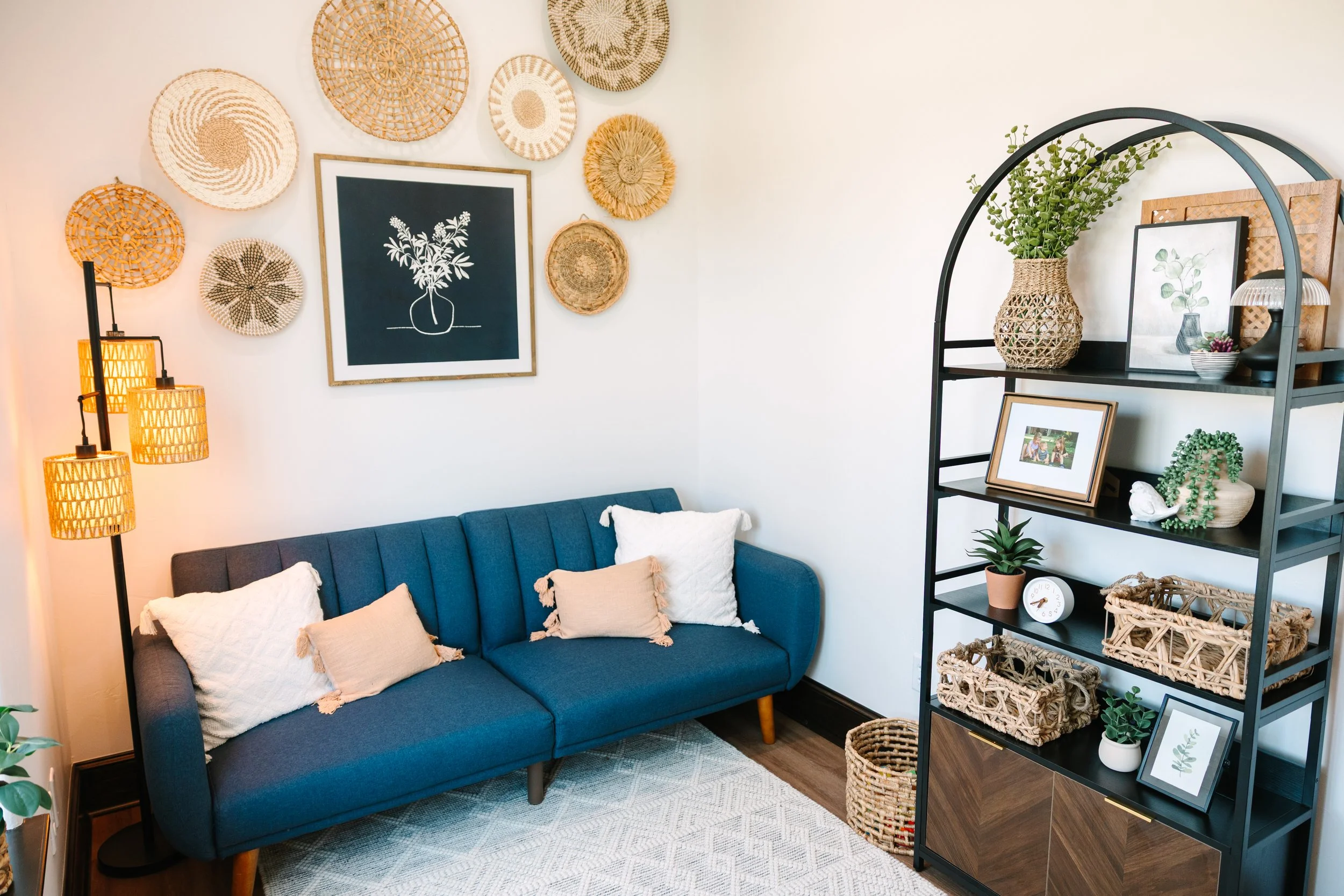 Living room with a blue sofa, beige and white pillows, a black shelving unit with decorative baskets, framed pictures, and plants, a wall decorated with woven baskets, and a multi-colored floor lamp.
