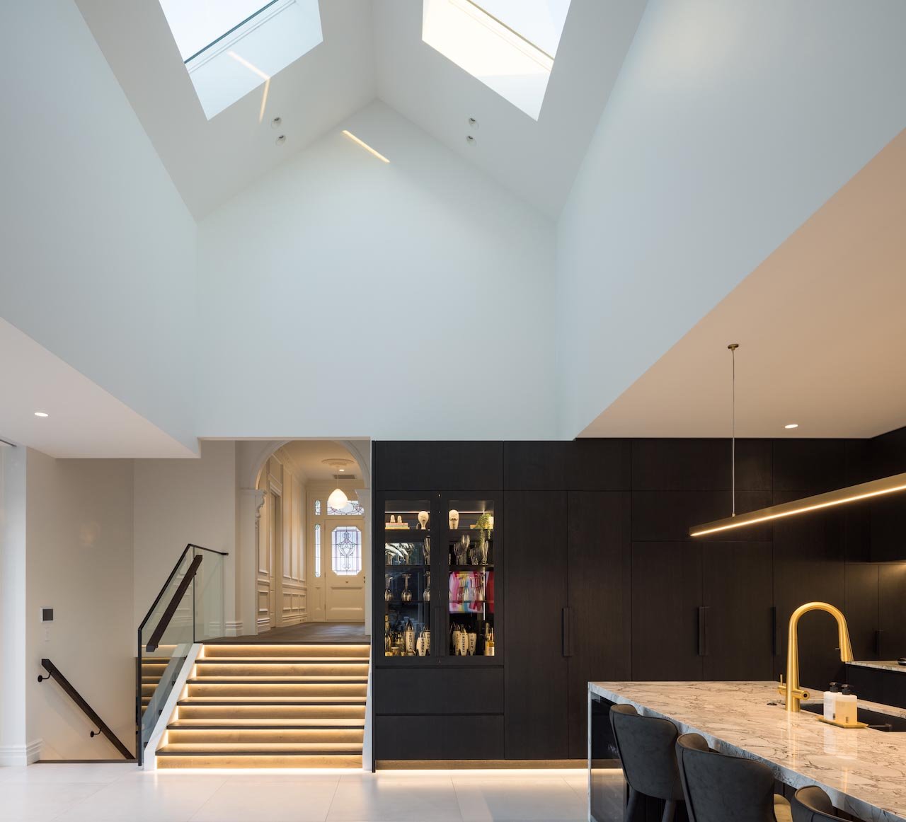 Zoomed out shot of dark kitchen in white room with very high ceiling and stairs to upper level of house.