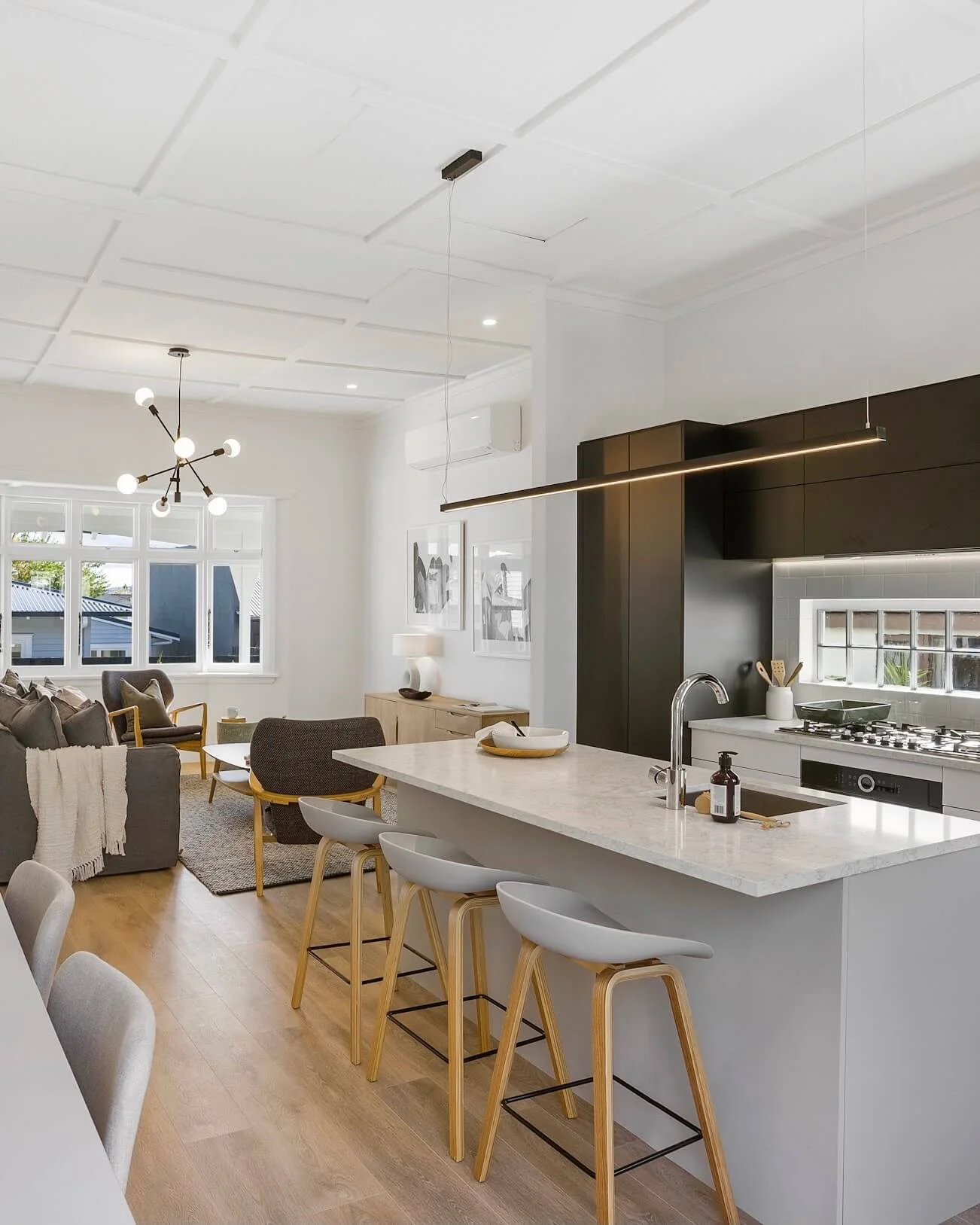 Modern open-plan kitchen and living room with a white marble island, gray and wooden bar stools, light wood flooring, minimalist decor, a sofa with throw blankets, and contemporary lighting fixtures.