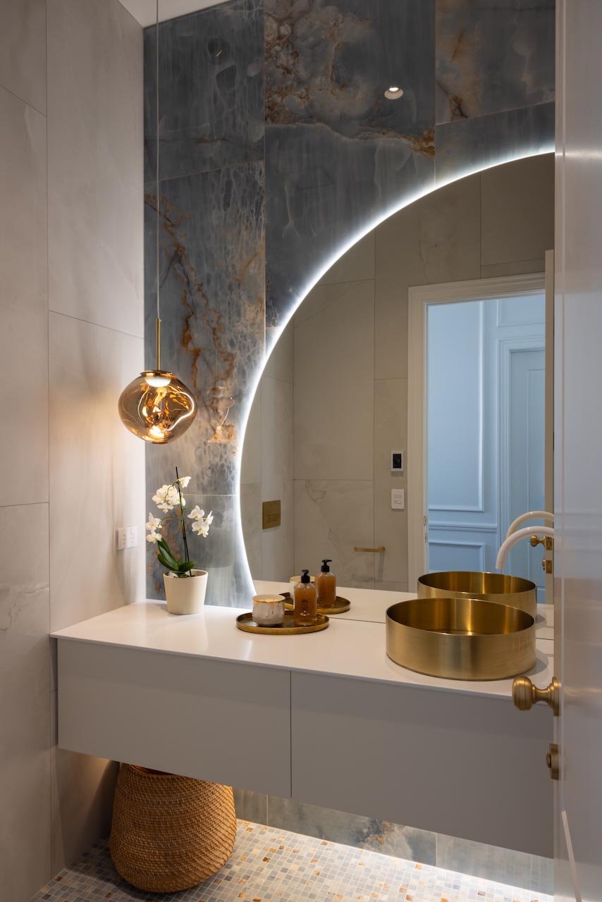 Powder room with round LED mirror and blue marble tiles with a brass basin.