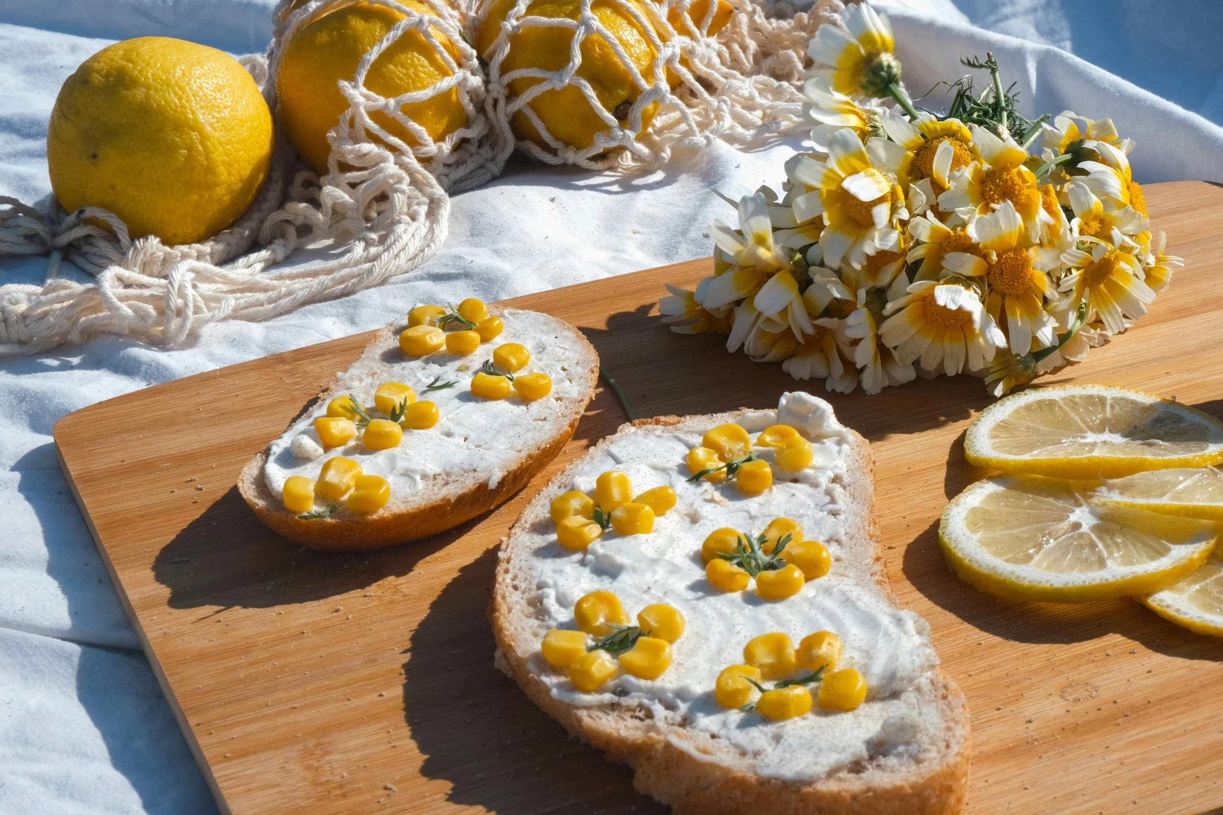 Two slices of bread with cream cheese and yellow berries on top, placed on a wooden cutting board. Nearby, there are lemon slices, a bunch of white and yellow daisies, and a couple of whole lemons wrapped in a netted bag, all on a white cloth.
