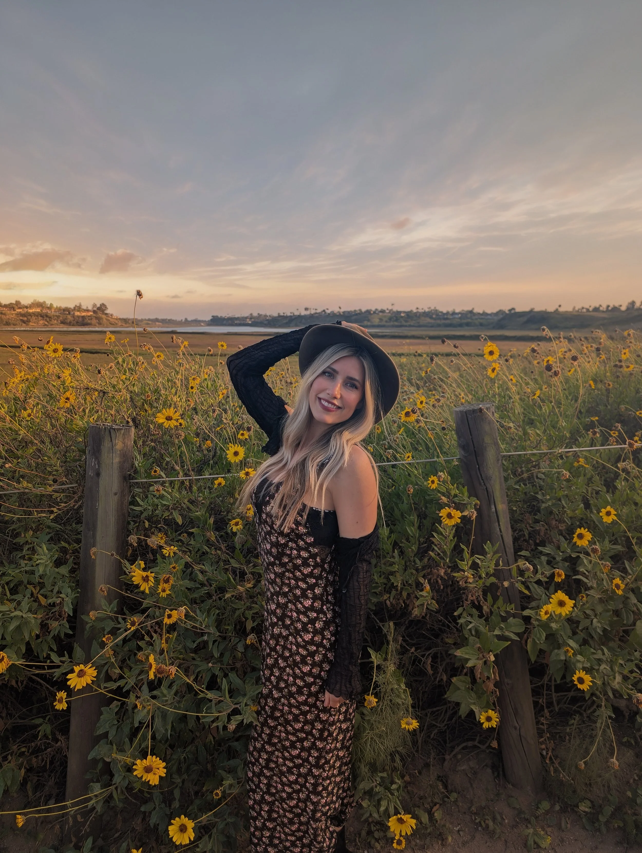 A young woman with long blonde hair, wearing a black hat, floral dress, and black lace sleeves, smiling while standing among yellow flowers in a field during sunset.