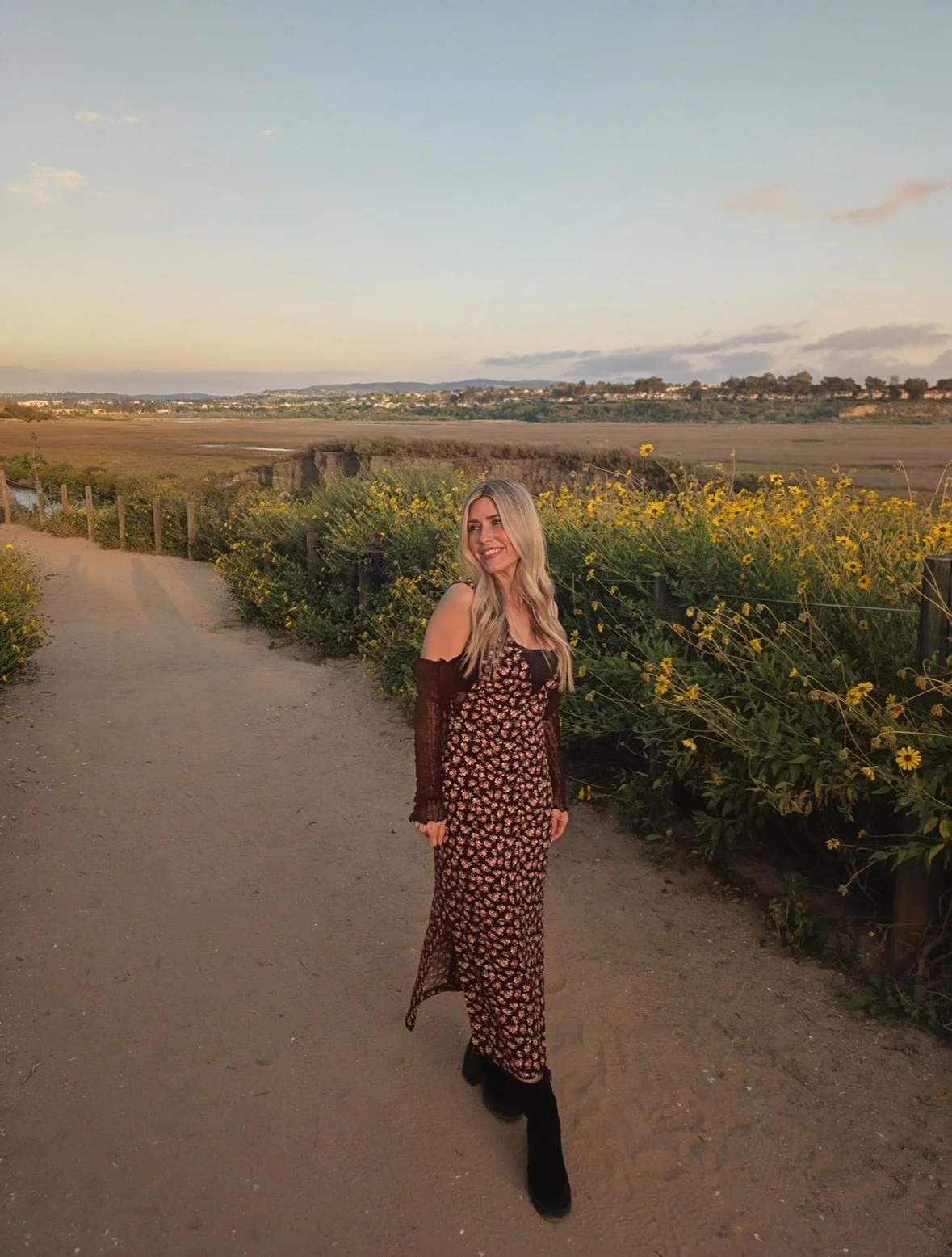 A woman walking along a dirt path lined with yellow flowers during sunset, with a landscape of open fields and distant hills in the background.