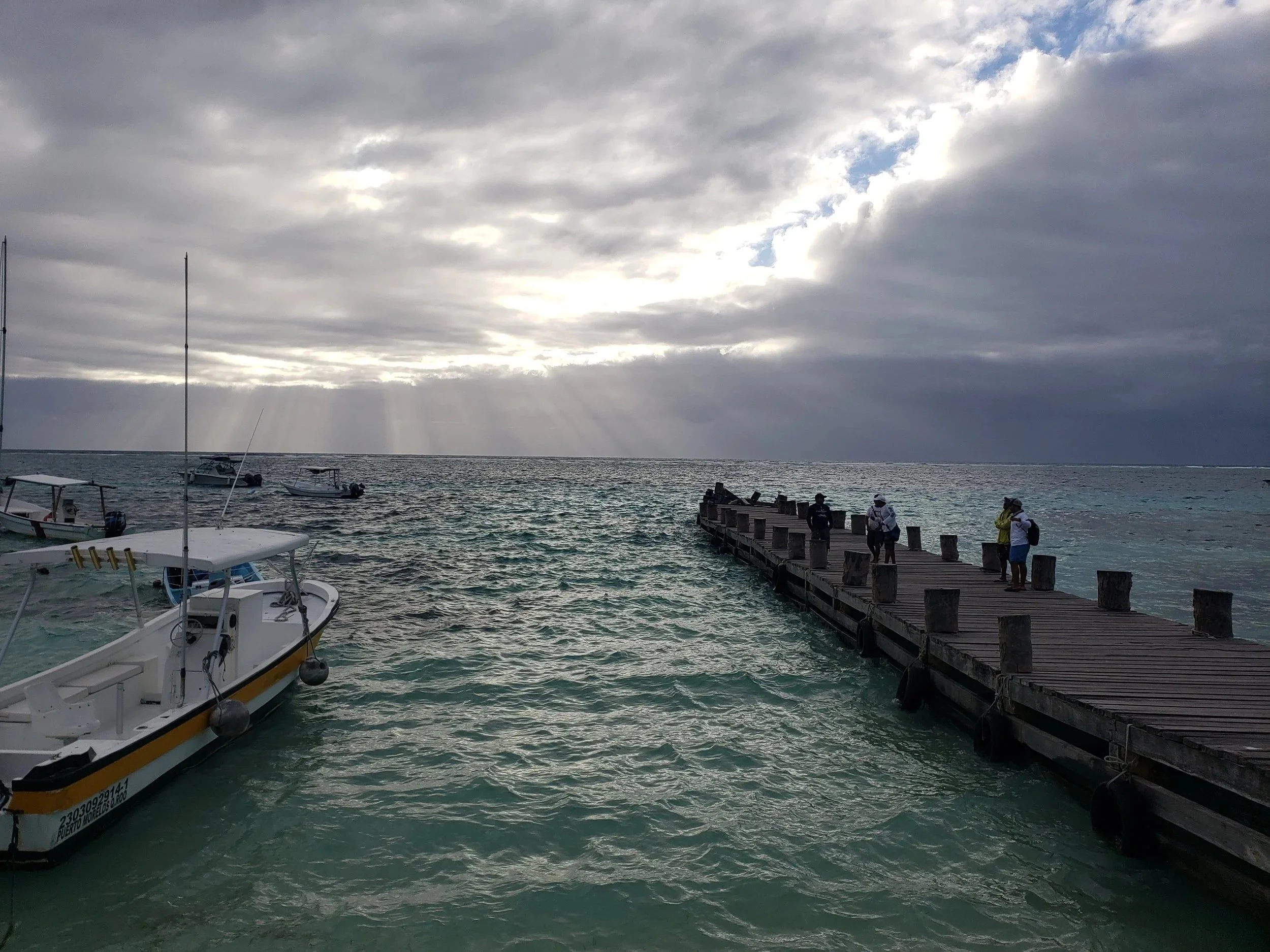 People standing on a wooden pier extending into the ocean under cloudy skies with rays of sunlight breaking through.