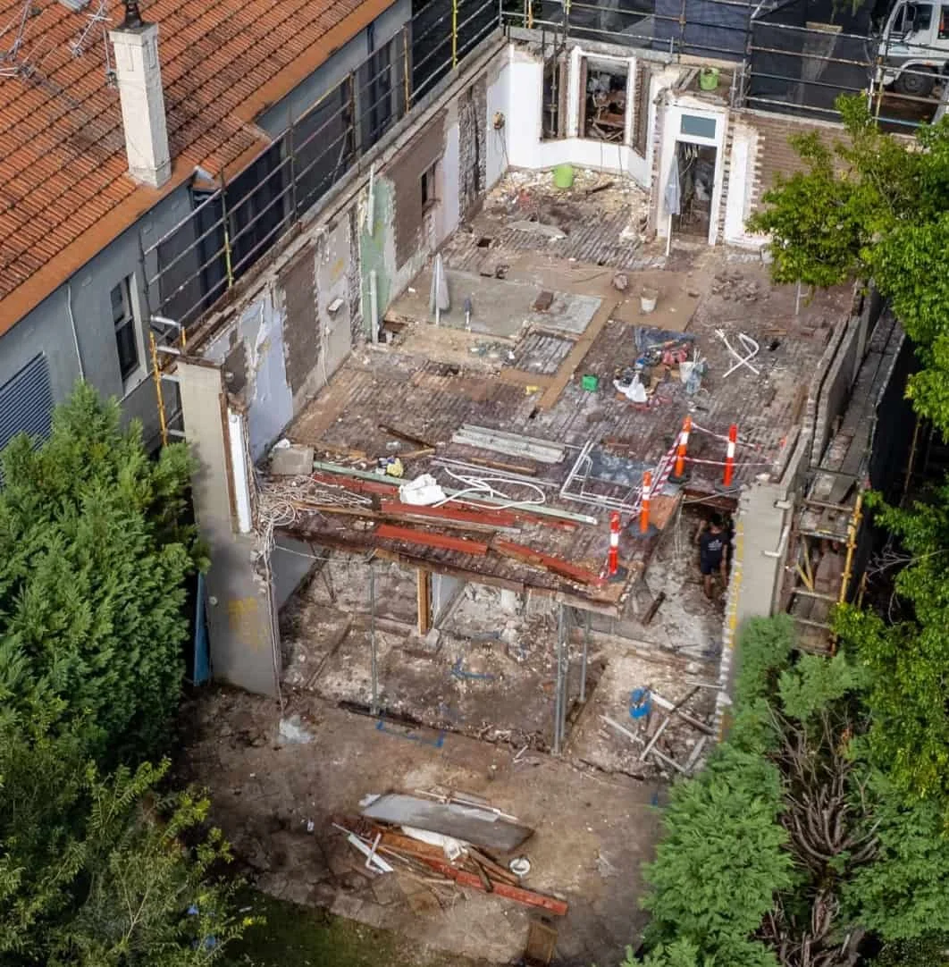 Aerial view of a building under renovation, with sections of the roof and walls torn down, construction materials and debris scattered, orange safety cones, and a worker.