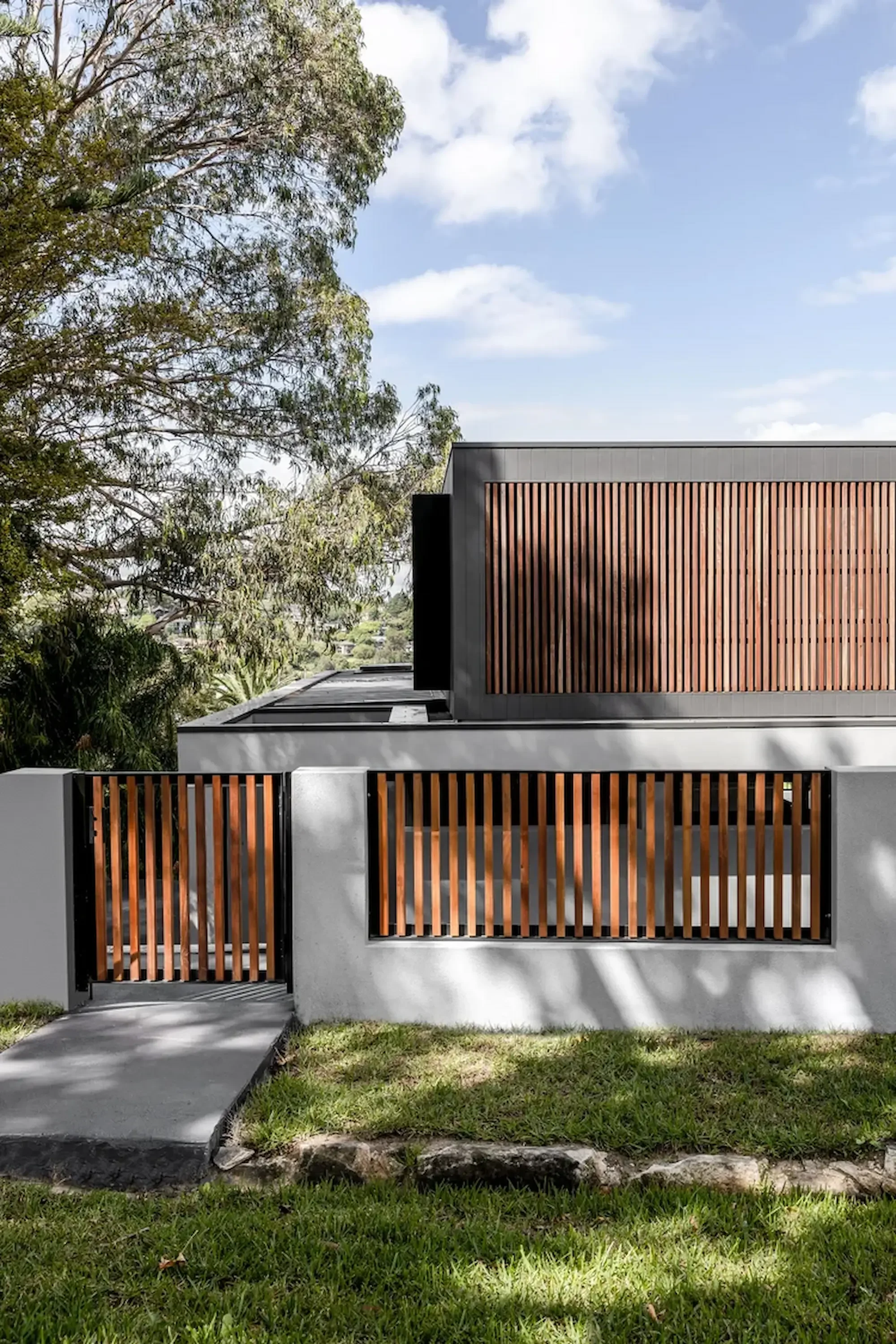 Modern house with gray exterior walls and wooden slat accents on the upper and lower sections, surrounded by a white concrete fence and gate, with green grass and trees in the background, under a partly cloudy sky.