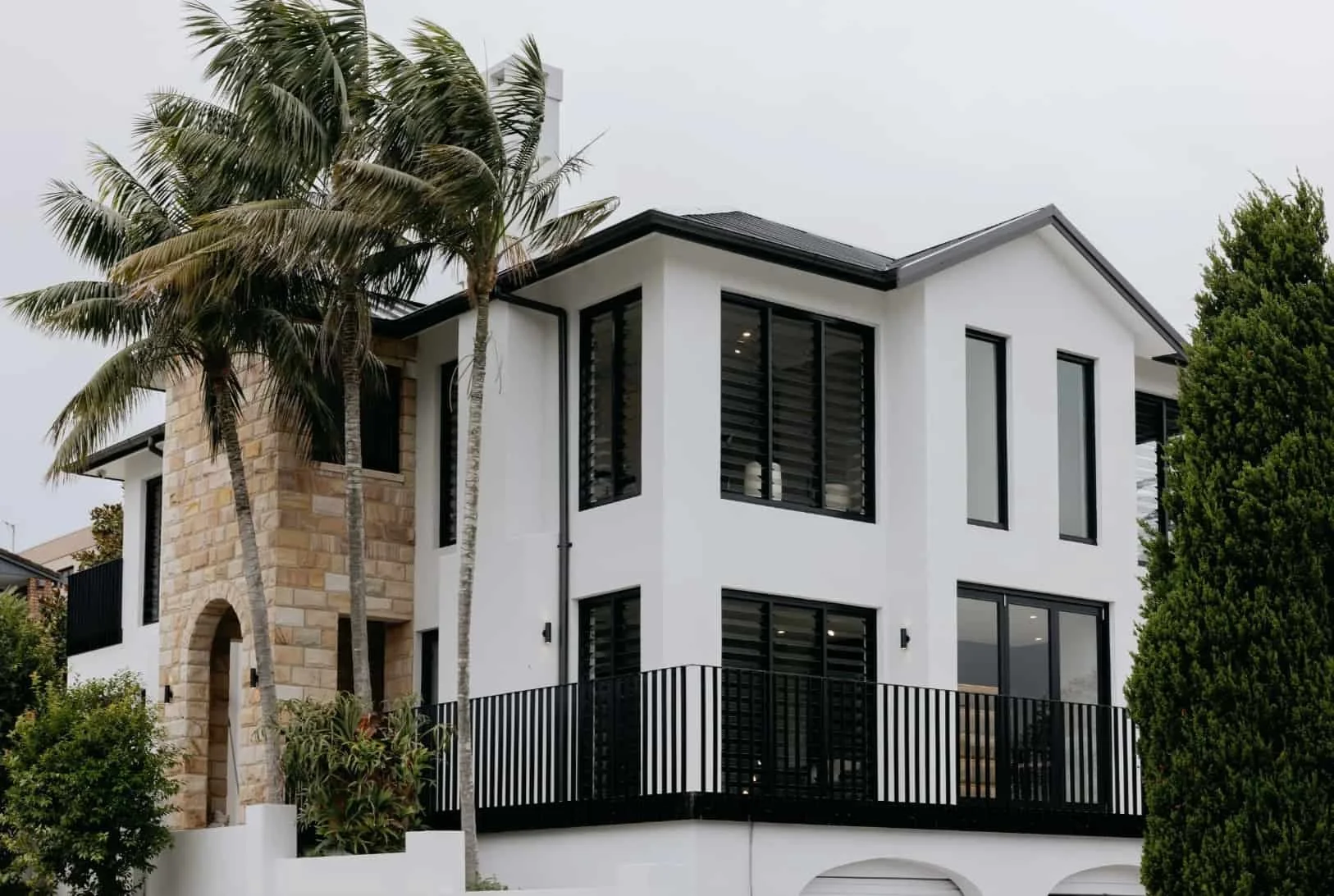 Modern two-story house with white walls, large black-framed windows, and a balcony with a black railing, surrounded by palm trees and greenery.