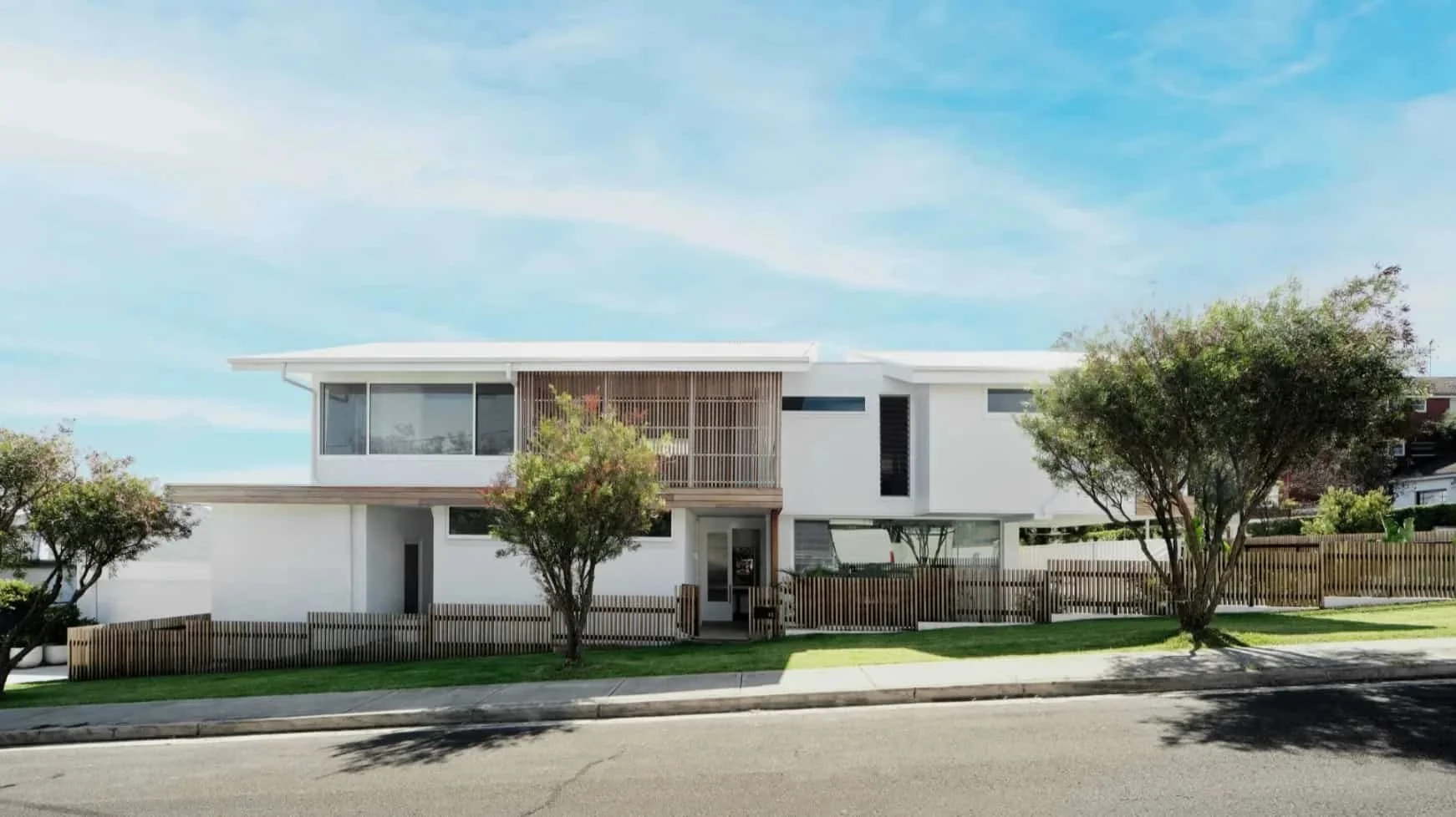 Modern white house with large windows, wooden slats, and a landscaped yard with trees, on a bright day with a blue sky.