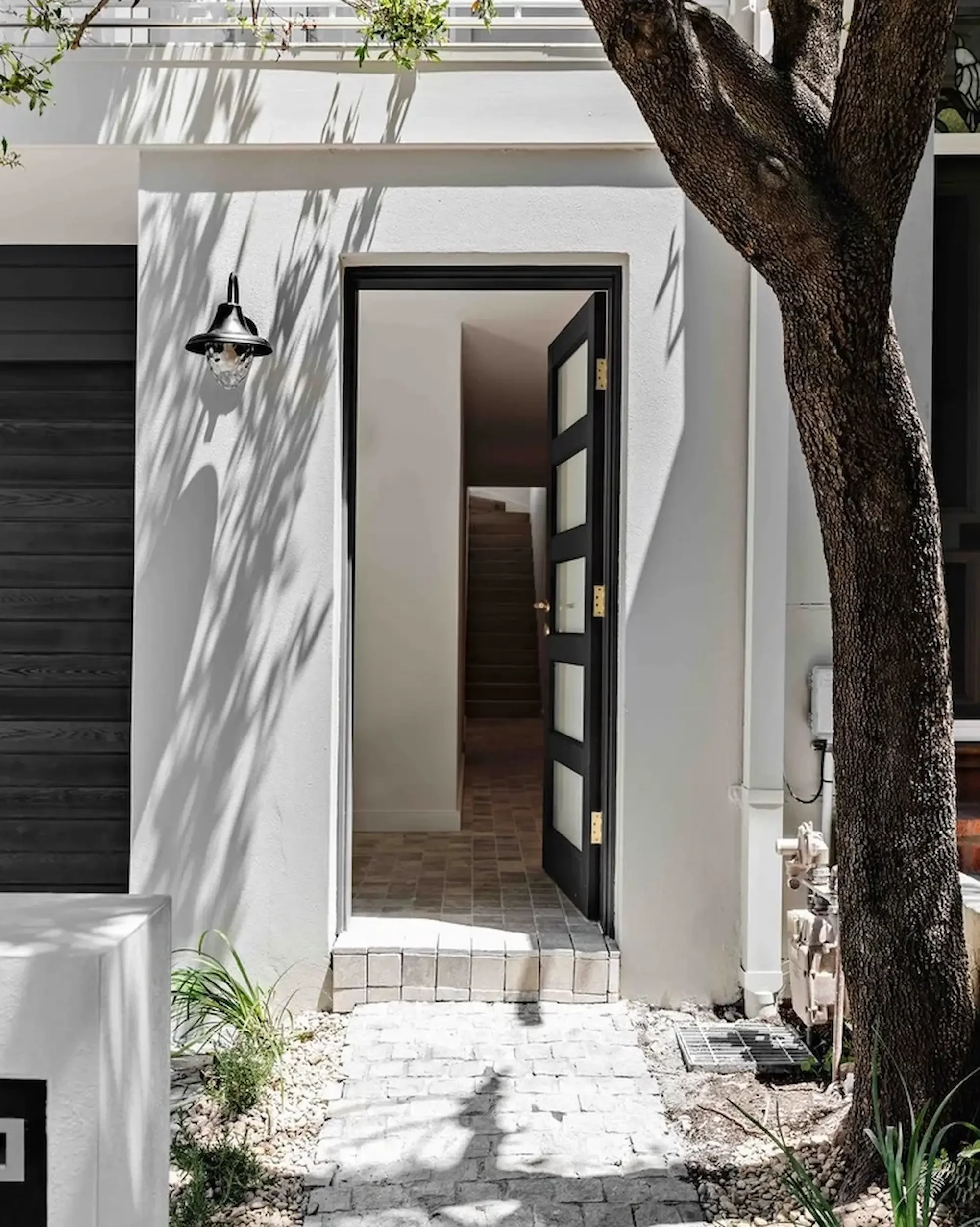 Open black door leading to interior staircase of a modern white house, with a tree casting shadows on the wall and a black outdoor light fixture.