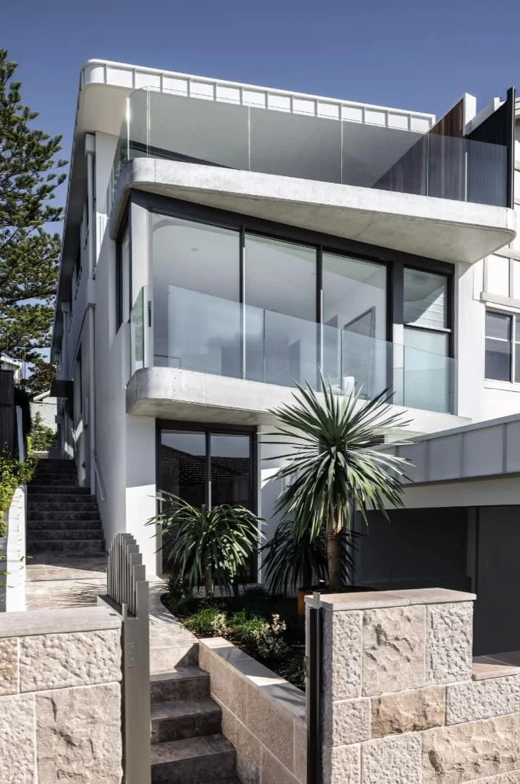 Modern multi-story house with large glass windows, concrete accents, and tropical plants outside, featuring a small front yard, stone steps, and an upper balcony with glass railing.