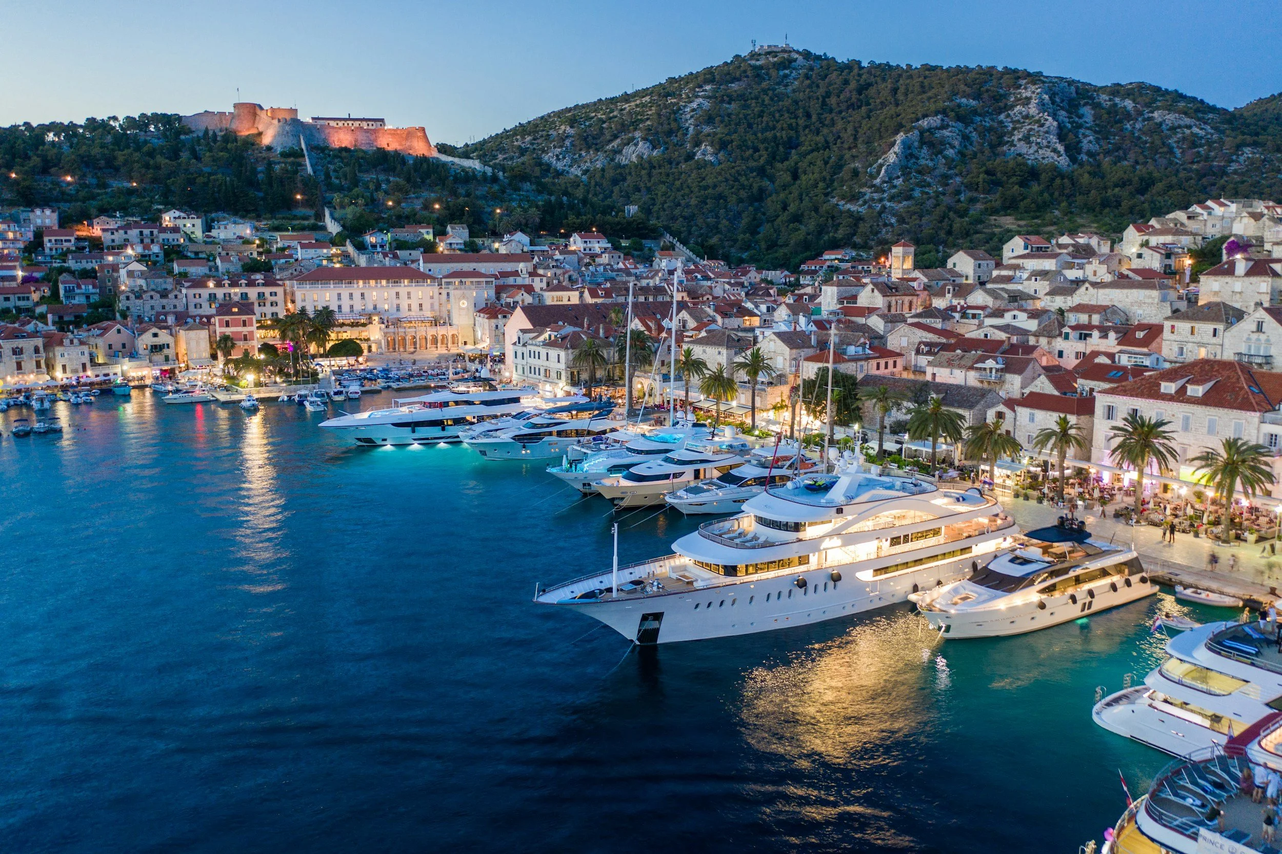 Night view of a harbor with luxury yachts docked along the waterfront, with a city with Mediterranean-style buildings and a hillside with a fortress and green hills in the background.