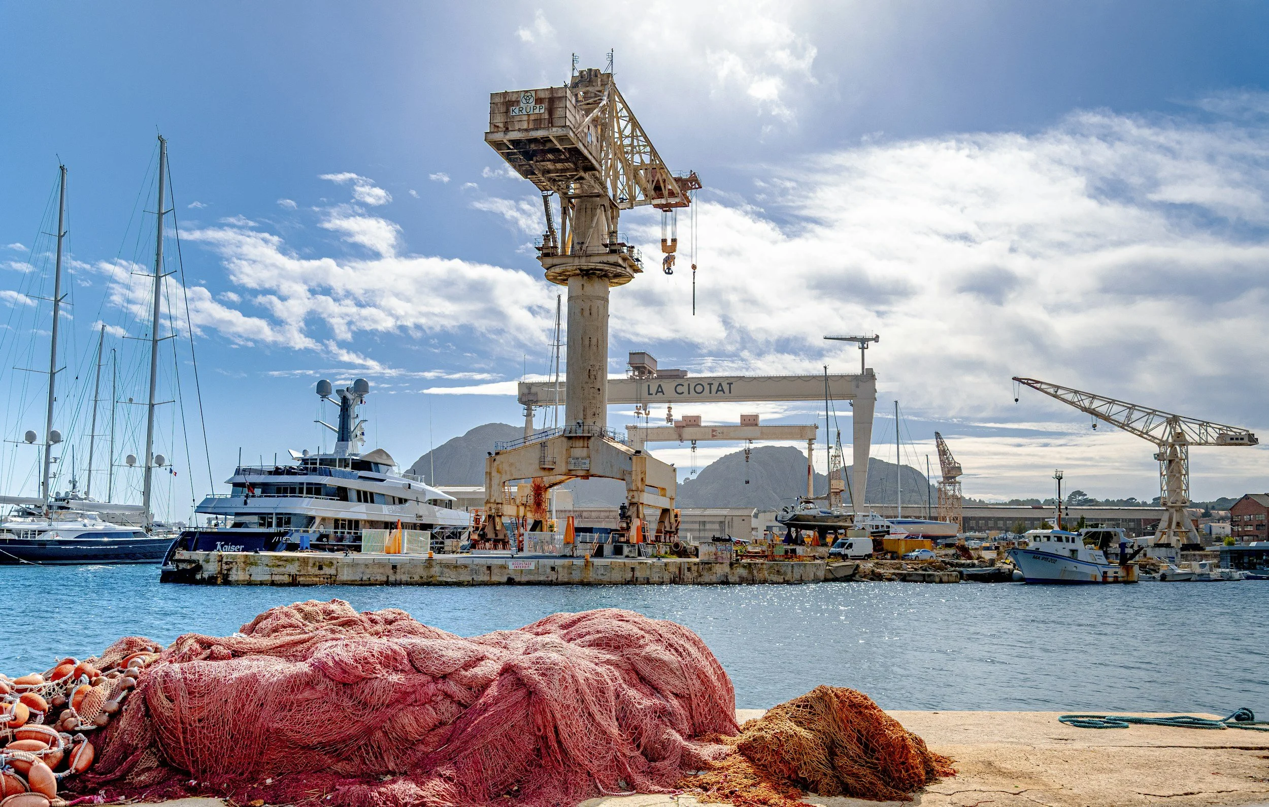 A harbor scene with fishing nets in the foreground, sailboats and yachts docked in the water, and large crane structures at the dock, with mountains and partly cloudy sky in the background.
