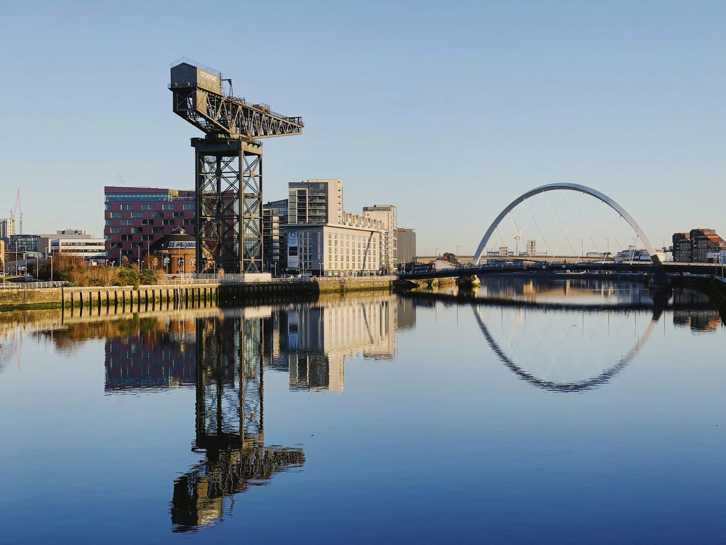 A cityscape with a river in the foreground reflecting the buildings, including a tall crane structure and a large white arch bridge.