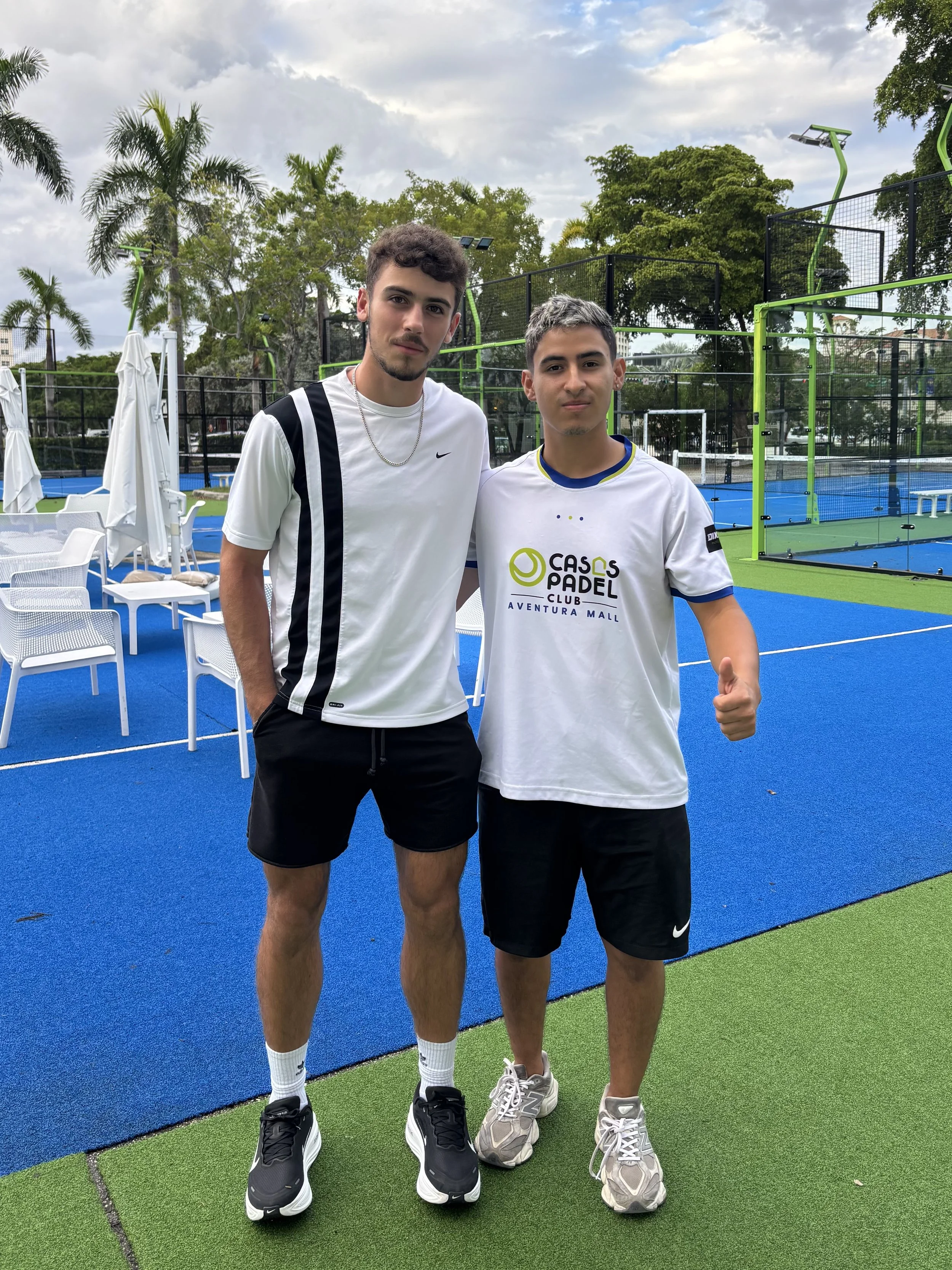The founder of BounceBackBalls and a man from Casas Padel standing in the academy. There are palm trees and a residential building in the background. 