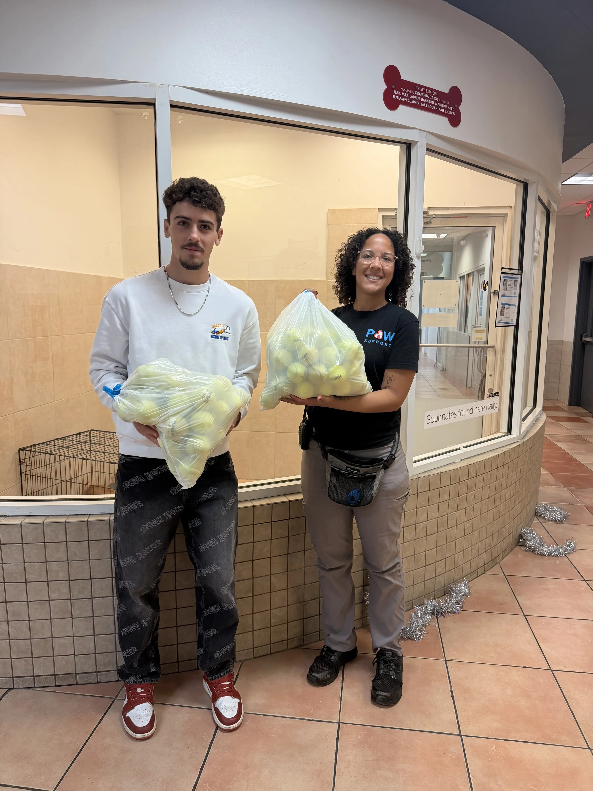 The founder of BounceBackBalls and a woman from the Humane Society of Greater Miami standing in the shelter, holding large bags filled with tennis balls, for BounceBackBalls.