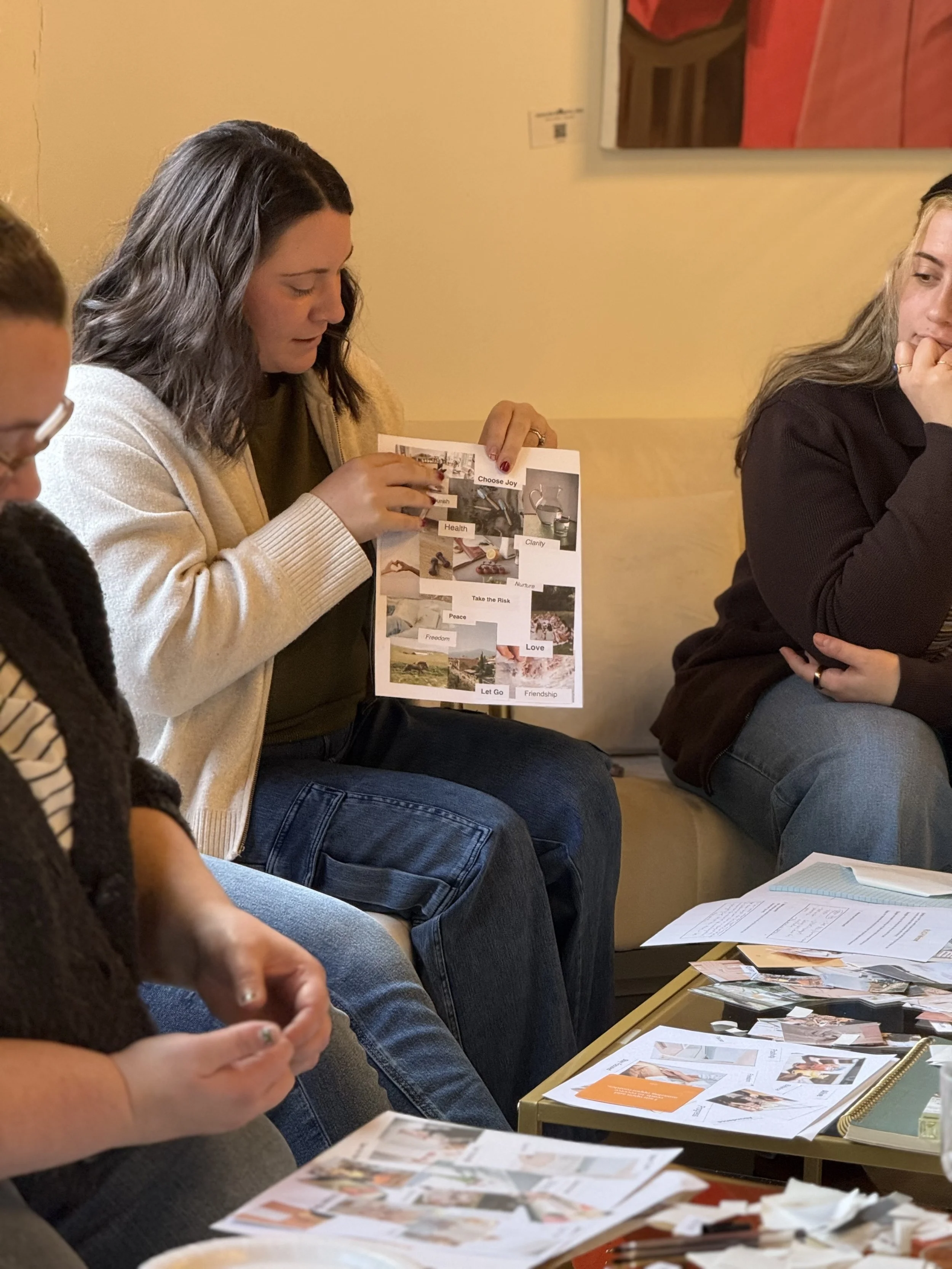 Three women seated on a couch, one holding a colorful collage with words like "Choose Joy," "Health," and "Love" on it, while the others look on and have papers and photographs on a table in front of them, suggesting a group discussion or planning session.