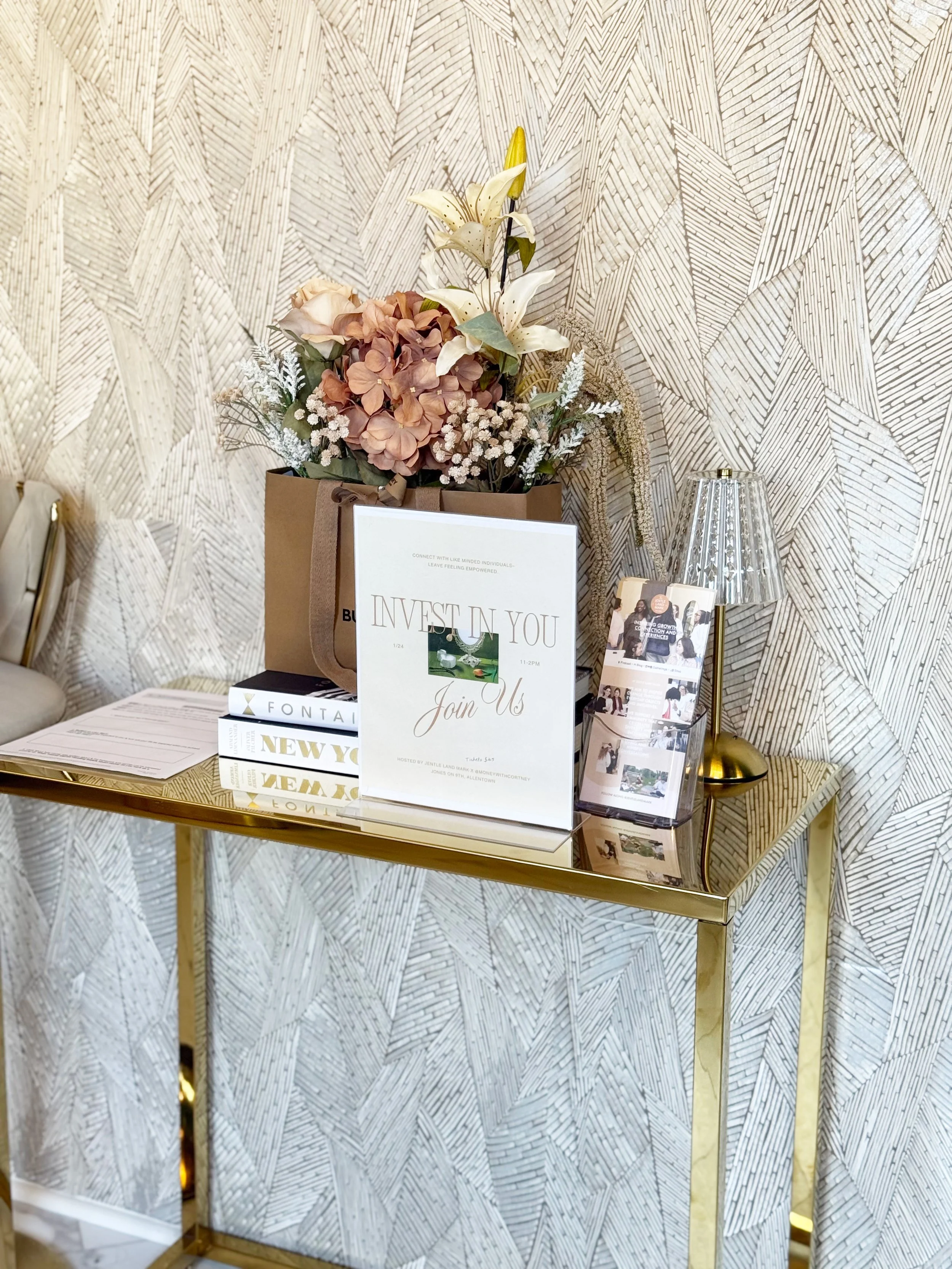 Decorative table with a flower arrangement, books, a sign for Jentle Land Mark's Invest in You event, a brochure stand against a textured wallpaper background at the Jones Collective in Allentown, PA, Lehigh Valley.