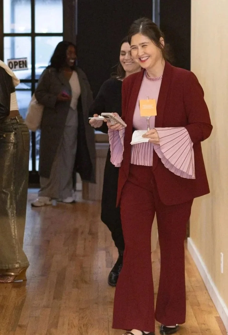 Women smiling and walking inside a building, one woman in a red suit and flared pants holding a glass and her phone.