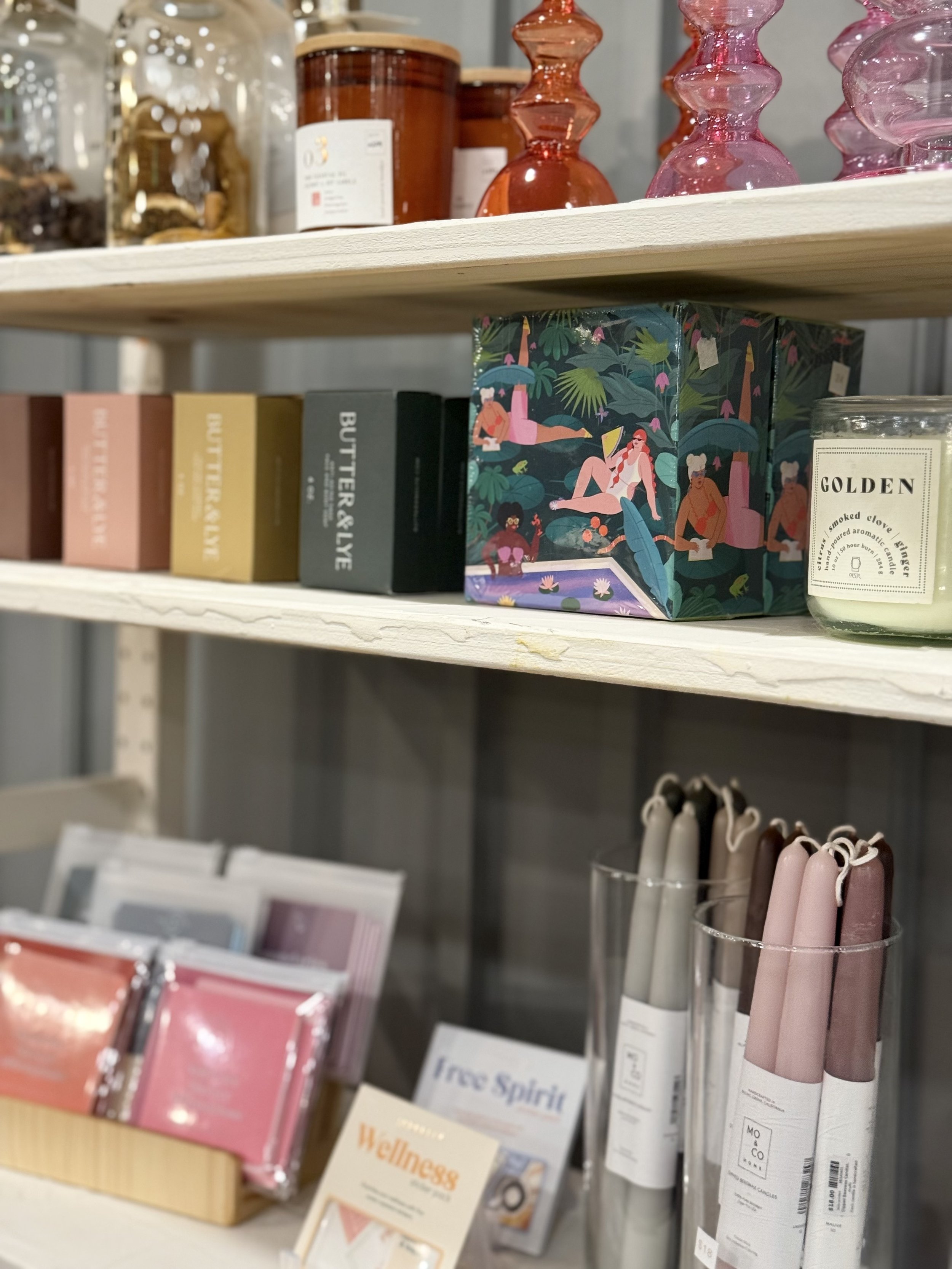 Shelves with candles, colorful boxes with artistic illustrations, skincare or wellness products, and pink and beige rolled candles.