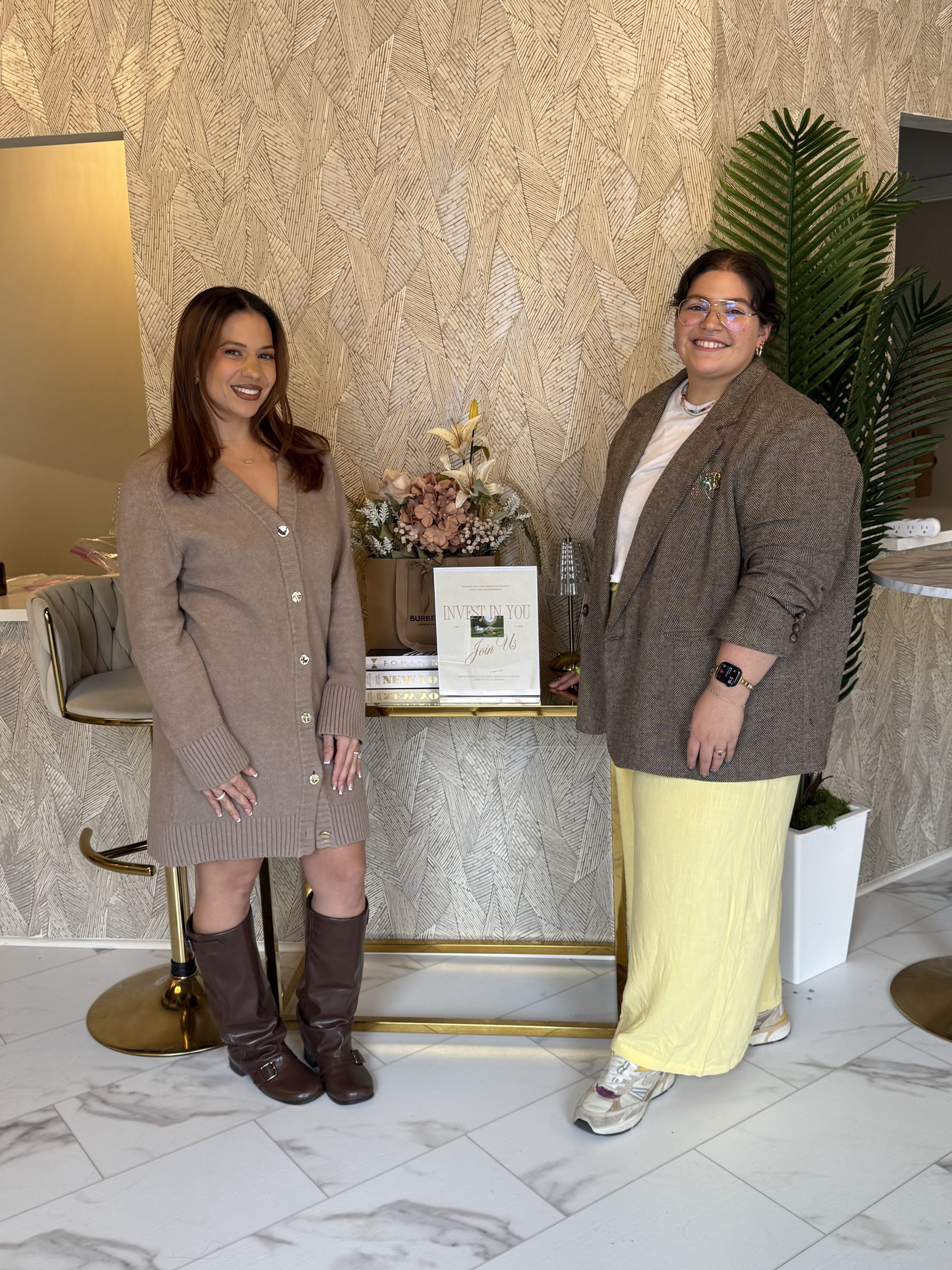 Two women, Cortney from Money with Cortney and Jenni, founder of Jentle Land Mark, standing inside a stylish room, smiling at the camera, with a decorative table with flowers and books between them, and a large green plant behind one woman.