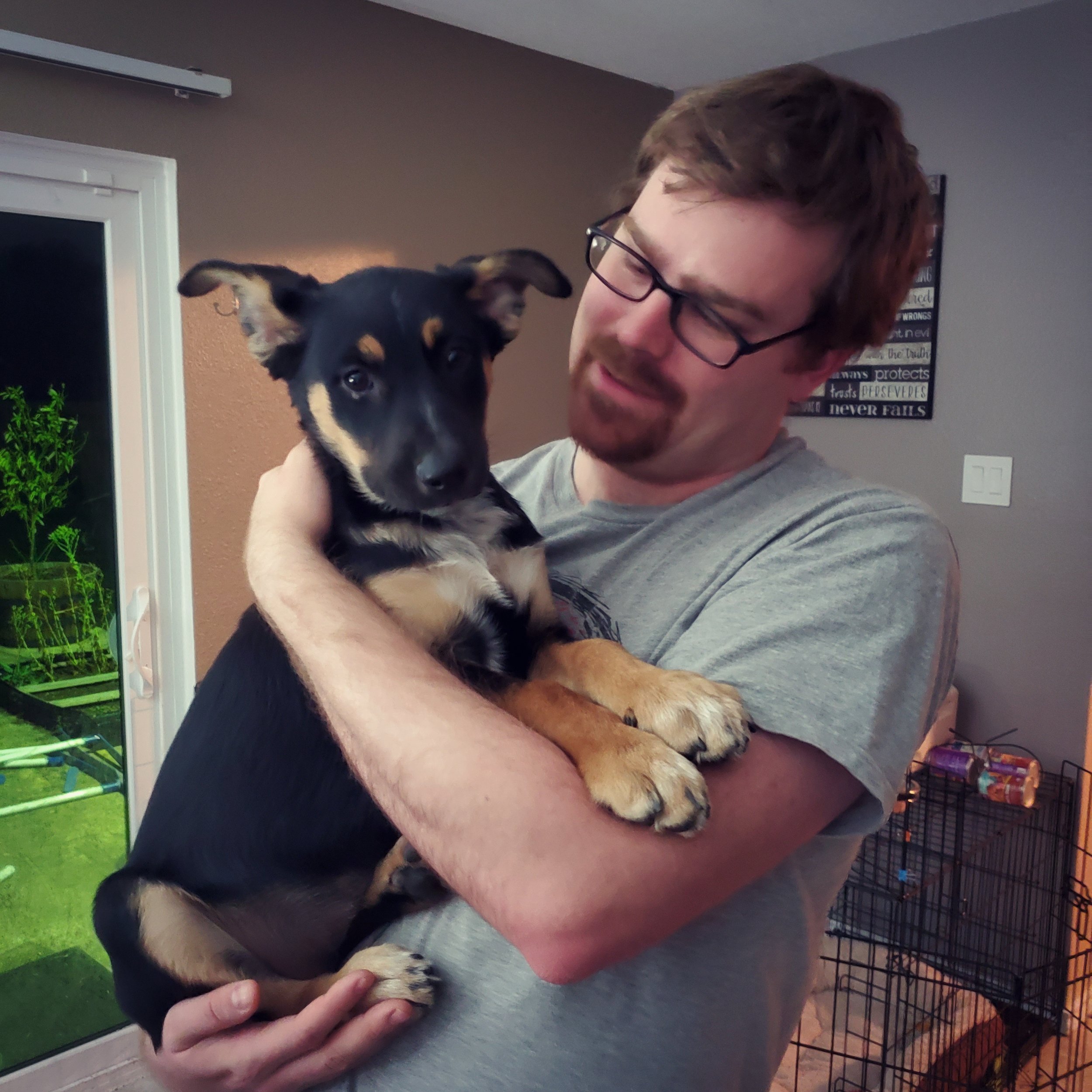 A man with glasses holding a young black and tan puppy inside a home.