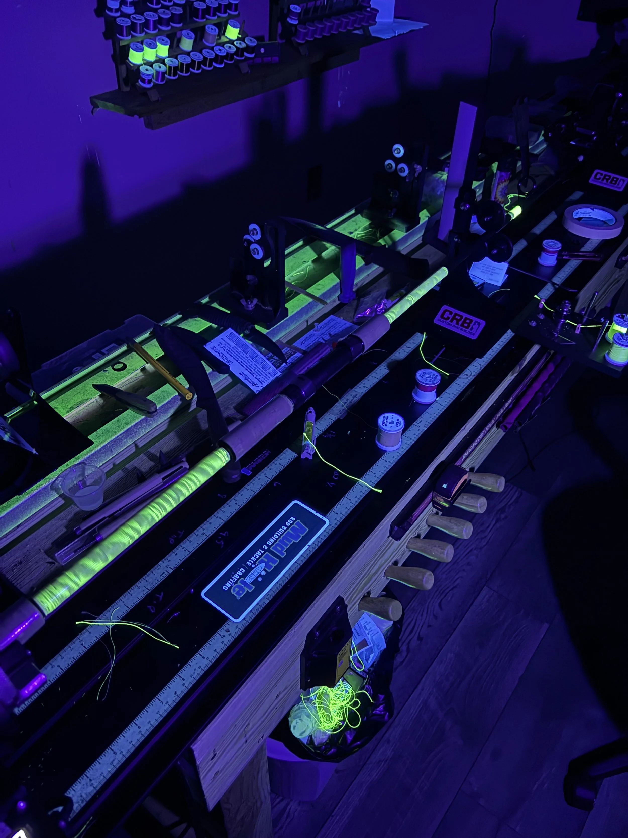 Fishing rod setup with glow-in-the-dark thread and reels, illuminated under blacklight on a workbench with measuring tape and tools.