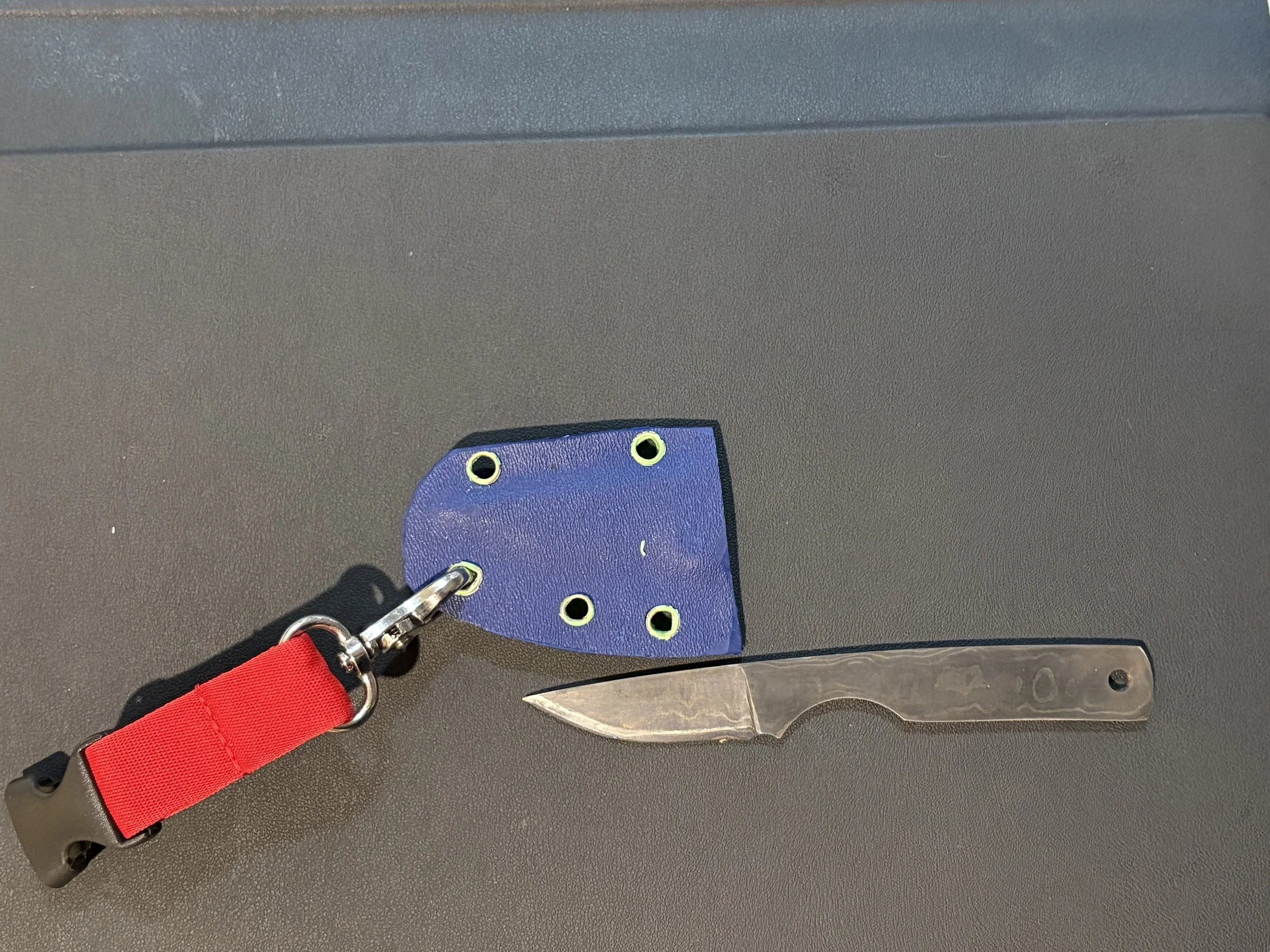 A small fixed-blade knife with a textured handle, a blue leather sheath with yellow holes, and a red strap attached to a keychain, all resting on a dark surface.