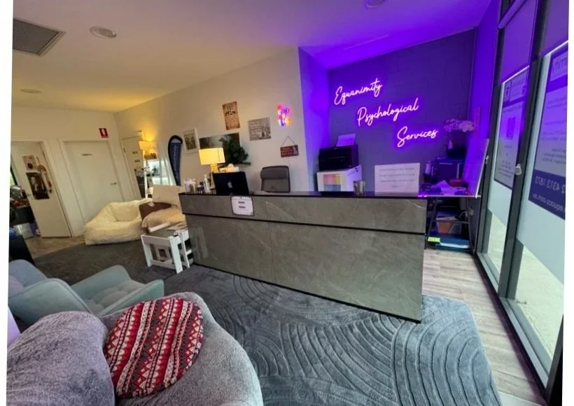 Reception area with a neon sign that reads 'Gynecology, Psychological Services' in purple on the wall. There is a reception desk, a waiting area with chairs and a sofa, and decorative items like plants and lamps.