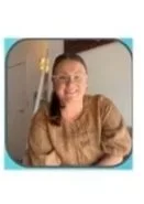 Smiling young woman with glasses in a brown top, sitting at a table, in a room with a light blue wall.