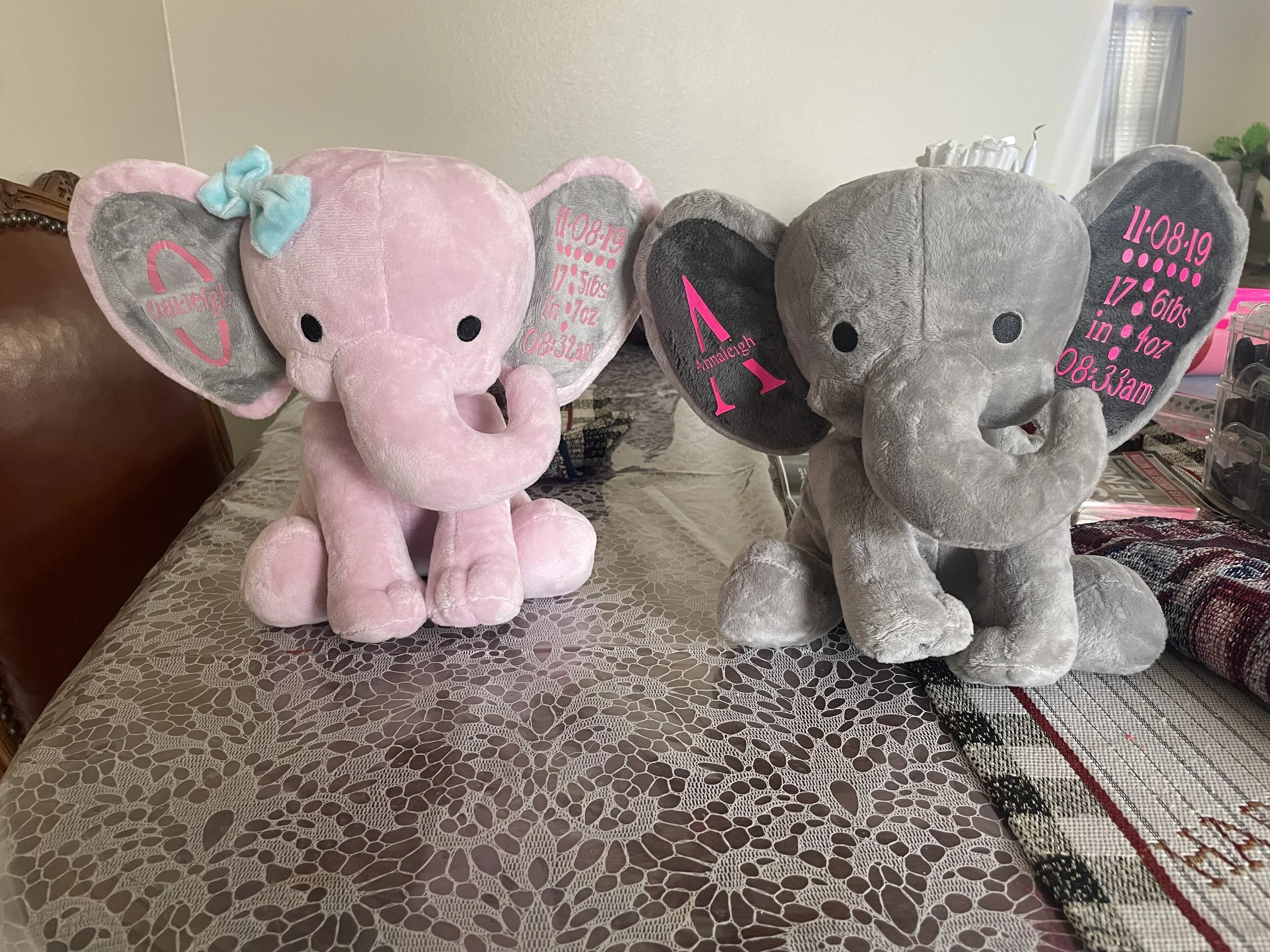 Two stuffed elephant toys on a lace-covered table. The left elephant is pink with a blue bow and has personalized text on its ear. The right elephant is gray with similar personalized text on its ear.