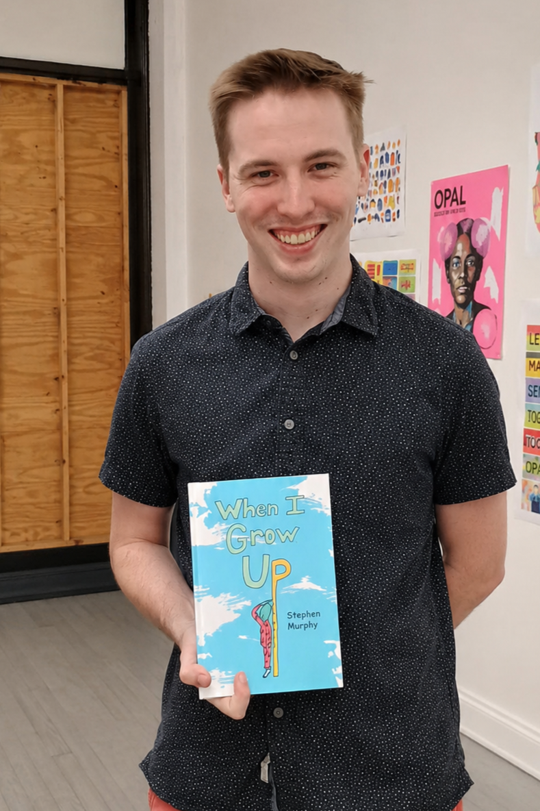A smiling young man with short light brown hair, wearing a dark, short-sleeved, button-up shirt with small white polka dots, holding a book titled 'When I Grow Up' by Stephen Murphy, standing in a room decorated with colorful artwork and posters on t