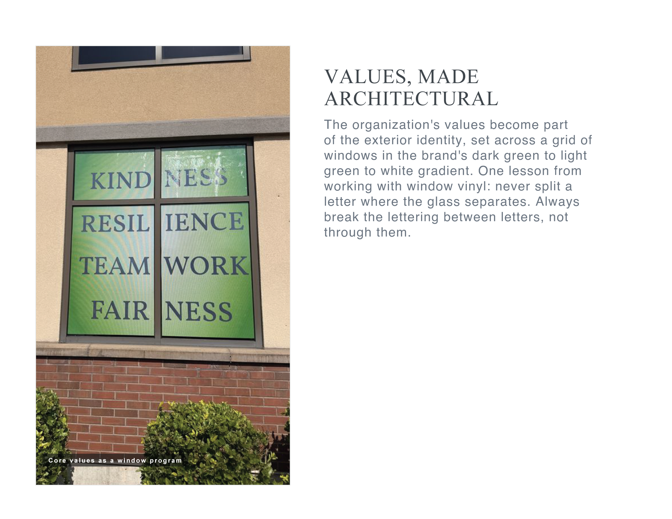 Window with four panes displaying the words 'KINDNESS', 'RESILIENCE', 'TEAMWORK', and 'FAIRNESS' against a green background, located on a building with beige exterior walls and brick lower section, surrounded by small green bushes.