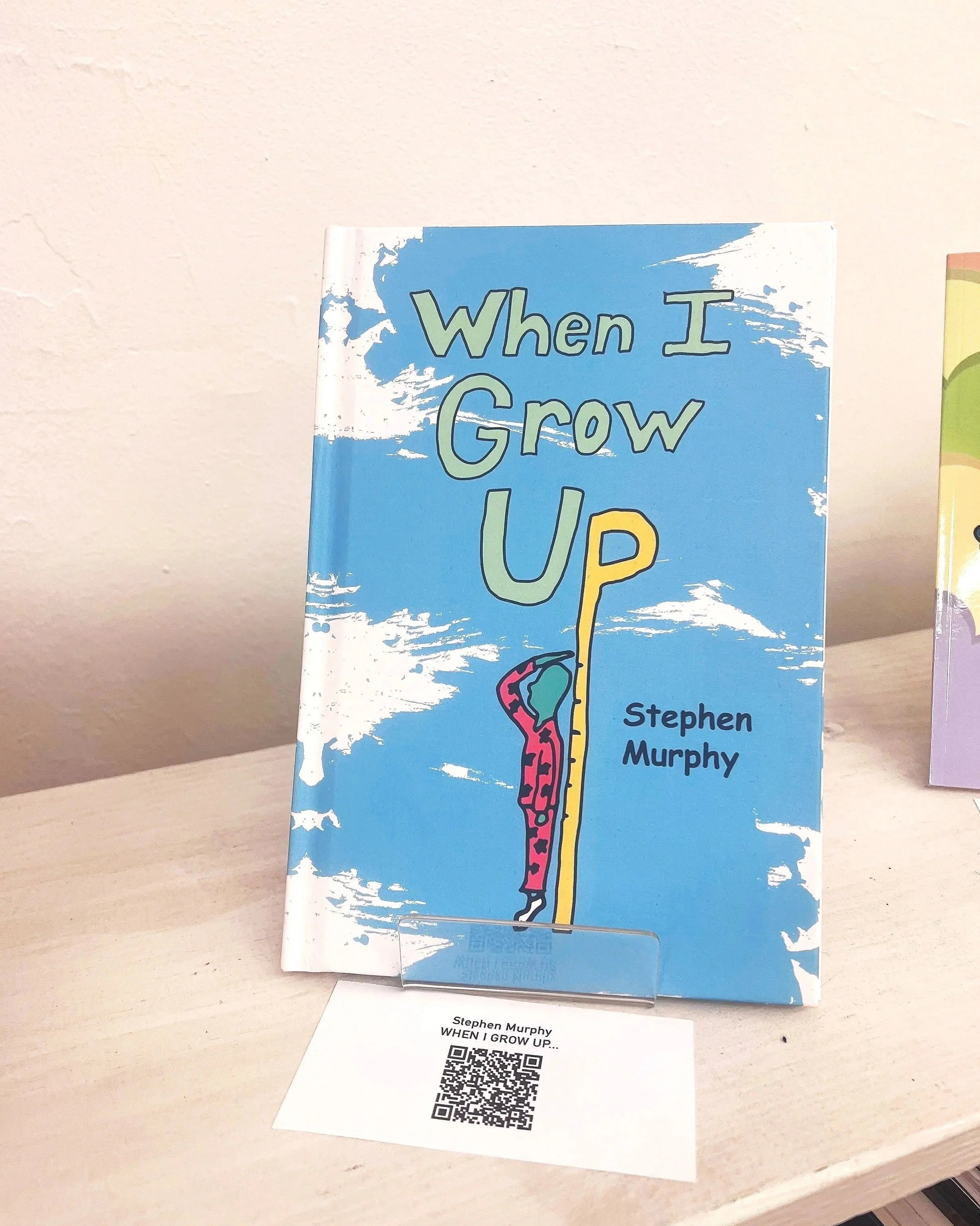 Book titled 'When I Grow Up' by Stephen Murphy standing upright on a table with a white wall background. A white paper with a QR code and the author's name and book title is lying in front of it.