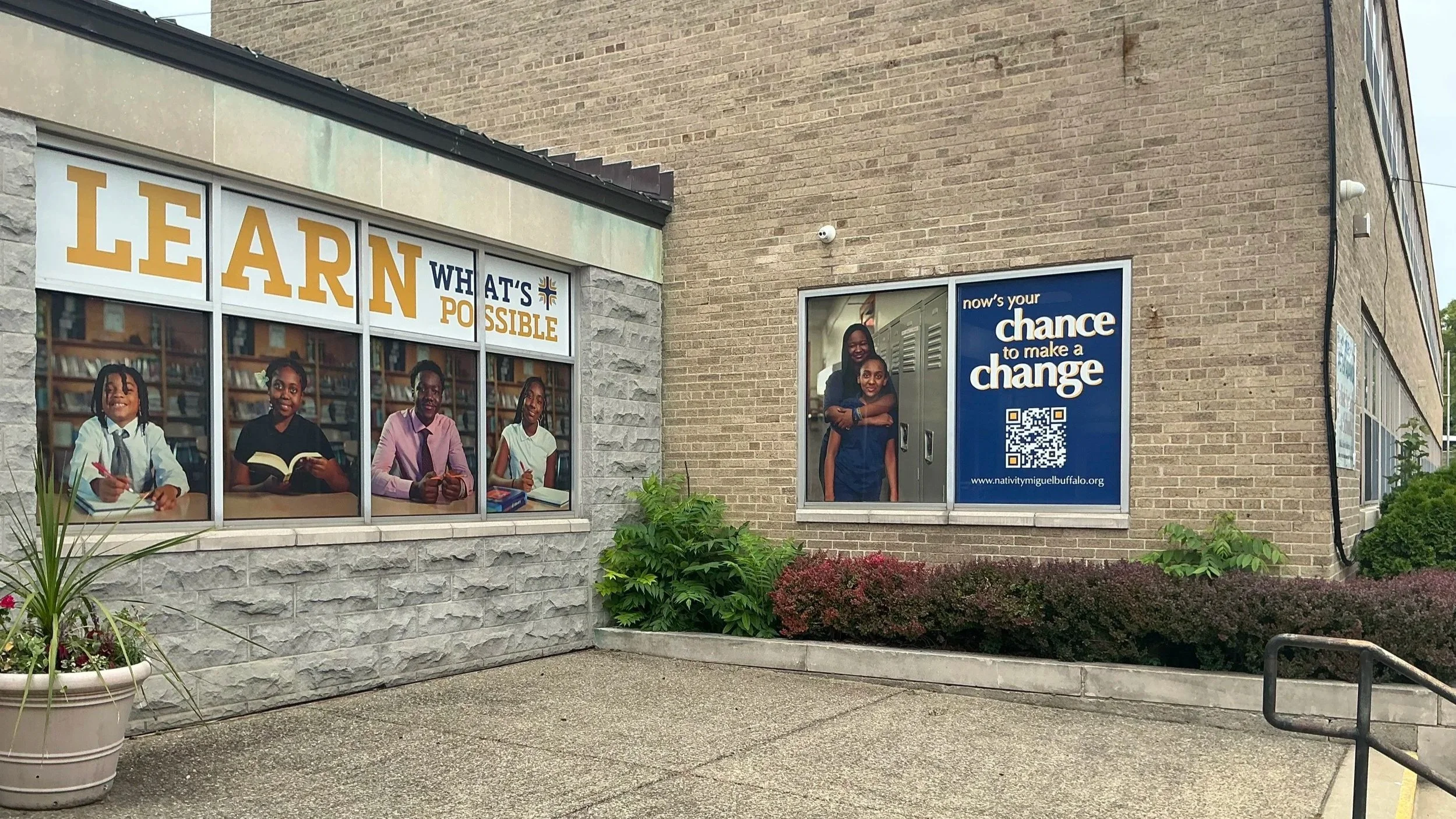 Exterior of a building featuring large posters with children and message promoting learning and making a change, including a QR code and website.