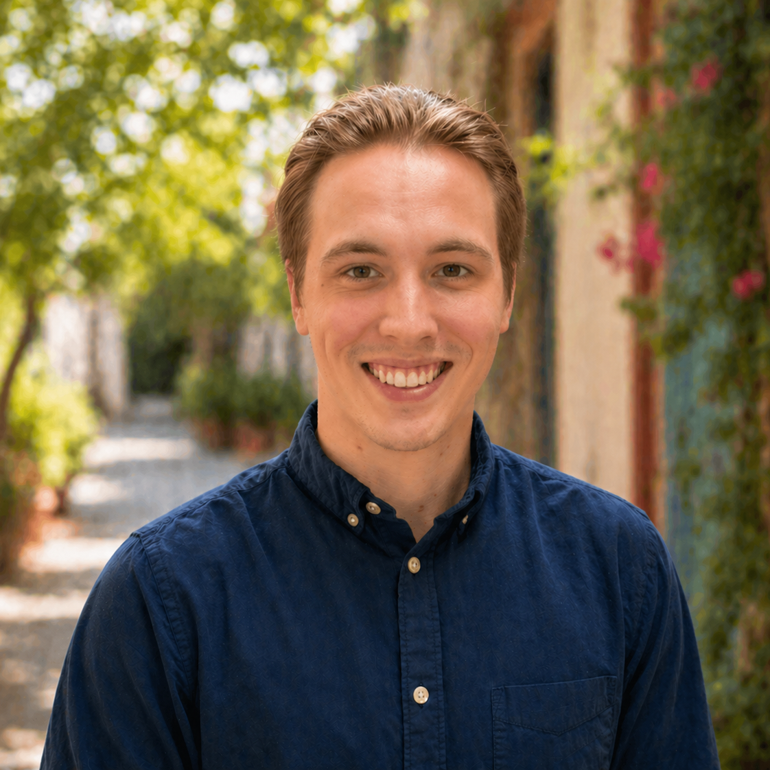 Young man with light brown hair and blue eyes smiling outdoors, wearing a navy blue button-up shirt, with a blurred background of trees and colorful building walls.