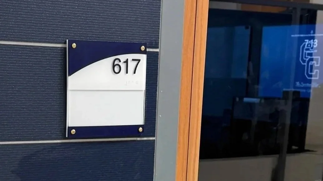 Close-up of a house number plaque displaying the number 617 on a dark-colored wall, with part of a door or window frame nearby.