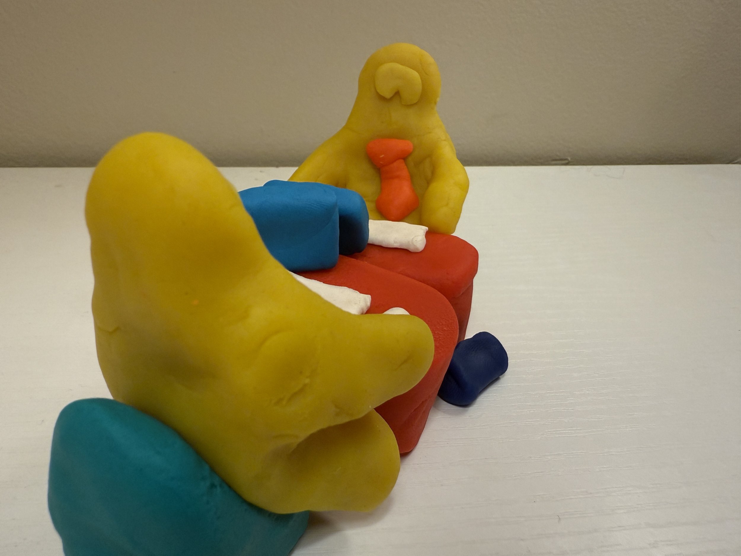 A colorful clay sculpture of an abstract figure with a yellow body, orange tie, blue and black accessories, sitting on a red surface, with a light gray background.