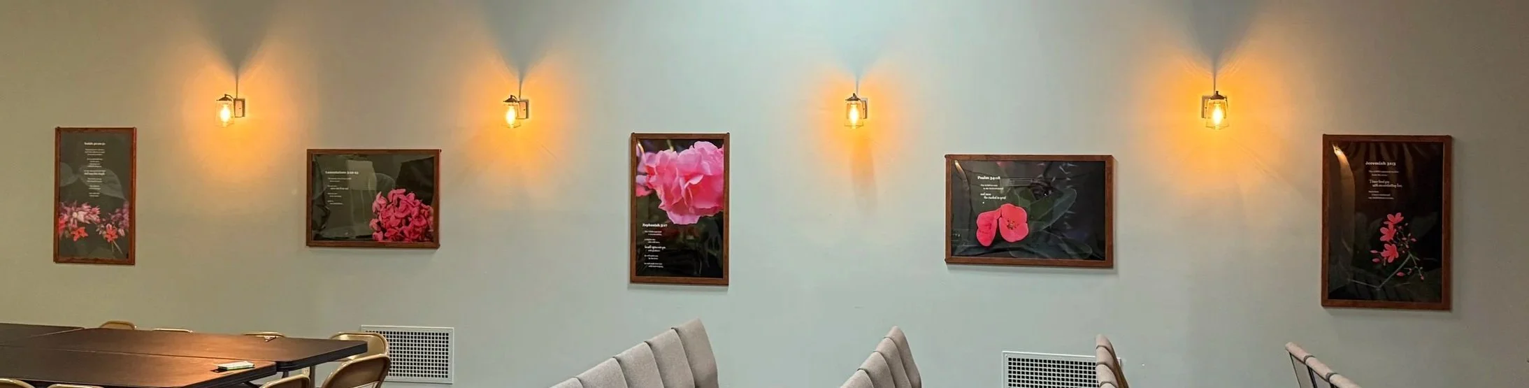 Six framed floral photographs with pink flowers are hung on a light-colored wall, accented by five wall-mounted light fixtures.