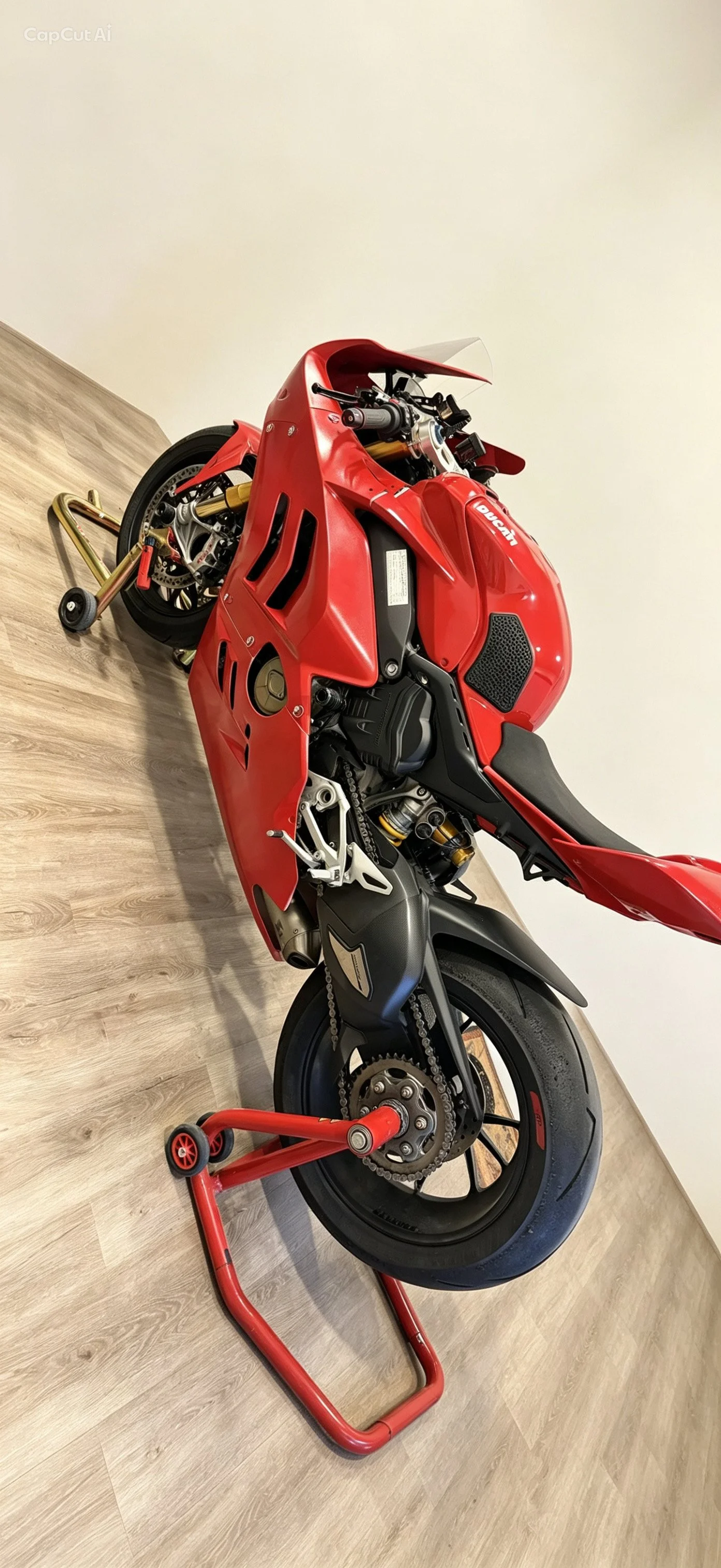 Red Ducati sport motorcycle on a motorcycle stand indoors, with a wooden floor and white wall.