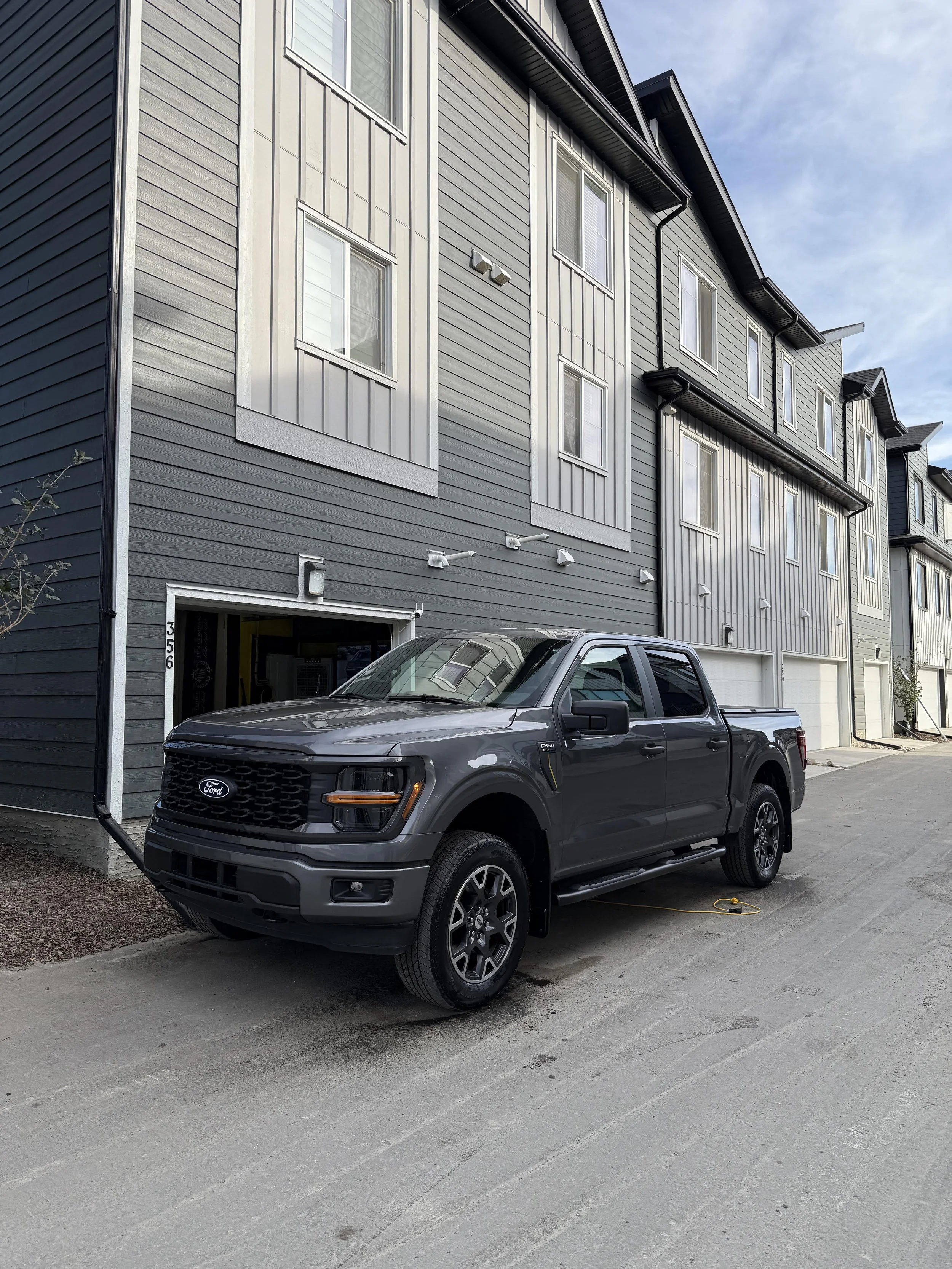 A black pickup truck parked in front of a modern apartment building with gray and white siding and multiple windows. The sky is partly cloudy.