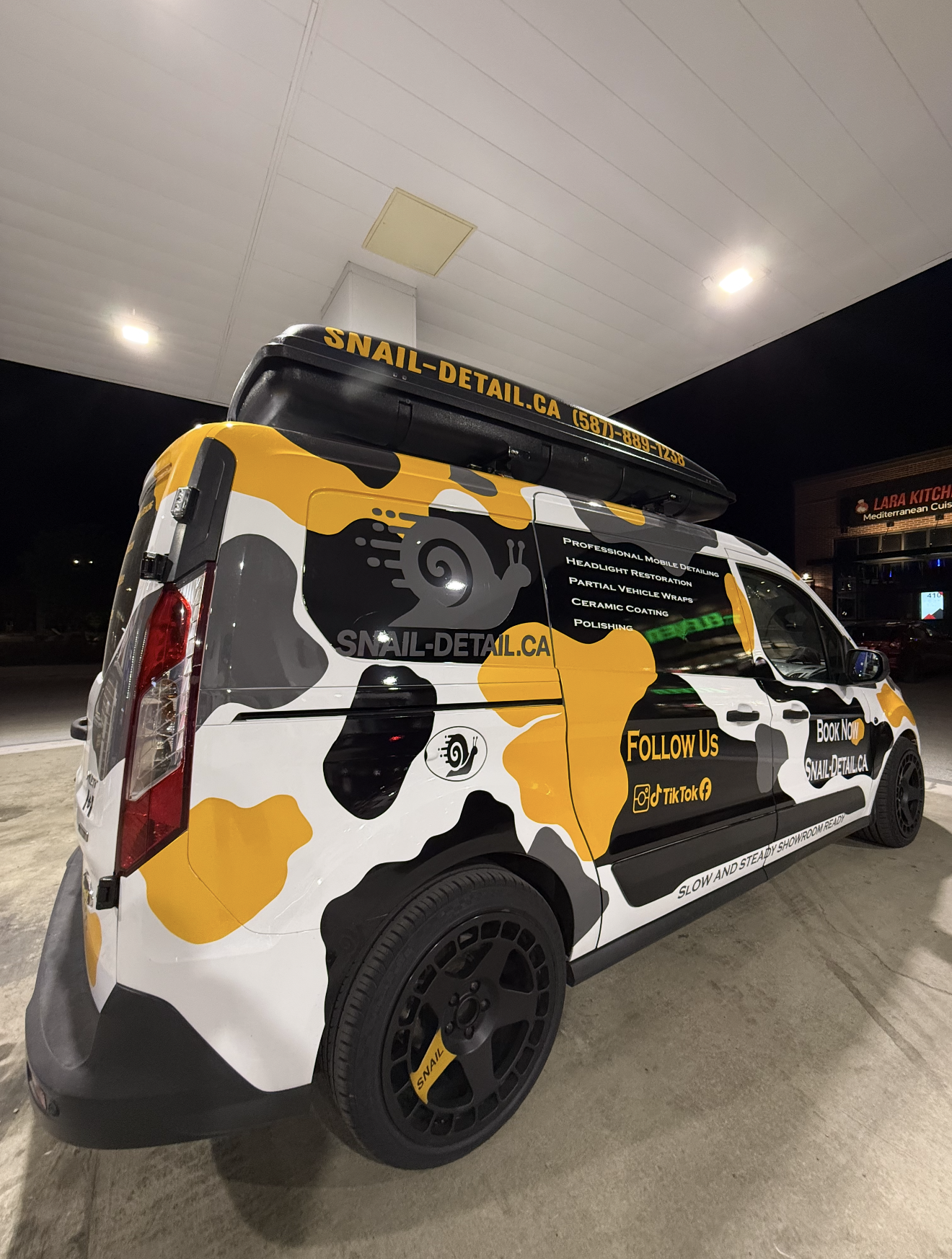 A mobile detailing service van with a yellow, black, and white camouflage design parked at night under a roof. The van displays the company's website, phone number, and social media icons, with text mentioning services like headlight restoration, ceramic coating, and polishing.