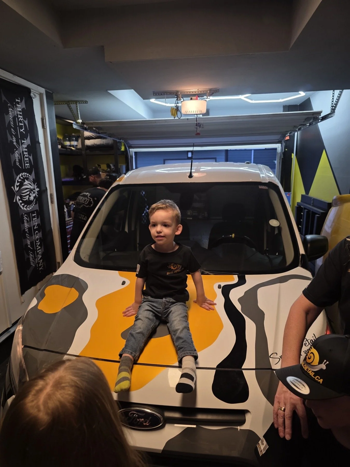 A young boy sitting on the hood of a vehicle painted with a tiger stripe pattern, inside a garage with children and adults around.