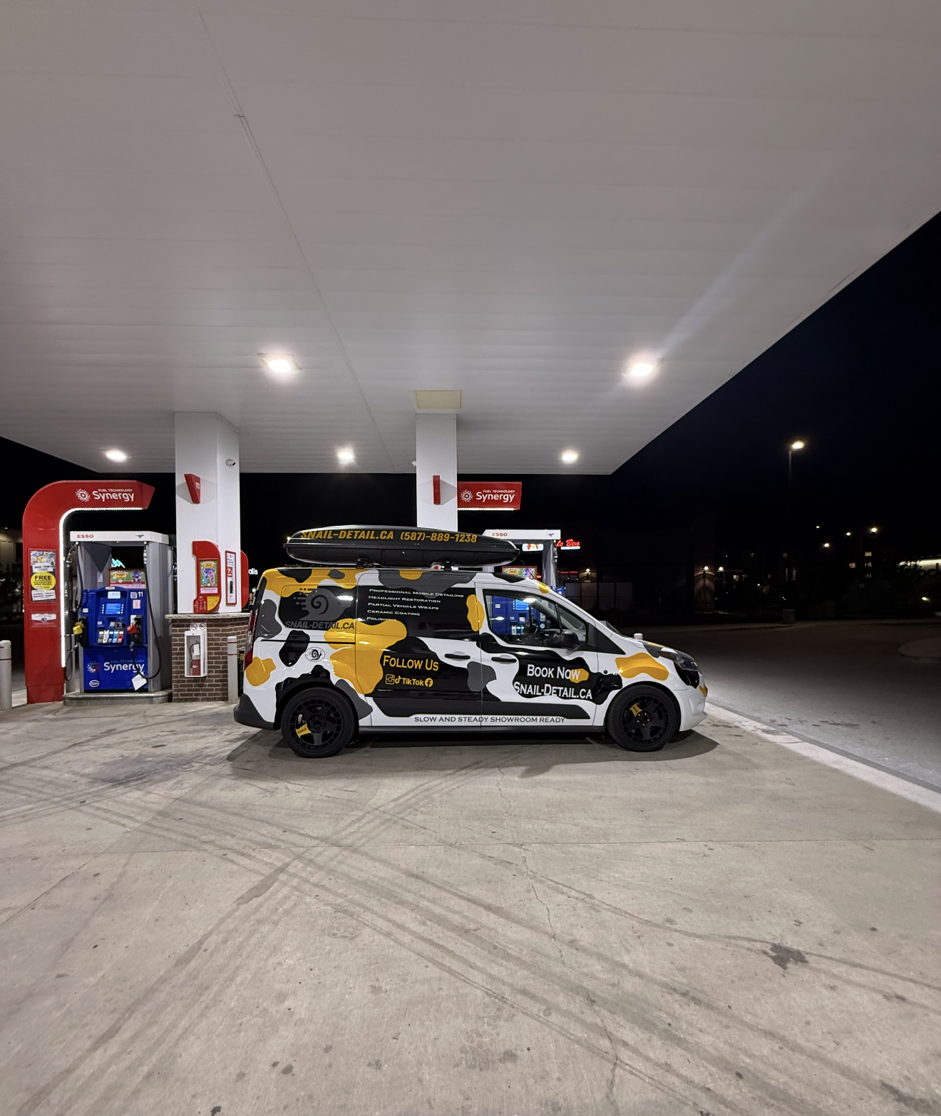A daytime photograph of a gas station with a black, white, and yellow advertising van parked near the pumps. The van displays the website 'SNAIL-DETAIL.CA' and promotes mobile detailing services. The gas station has two visible fuel pumps with the 'Synergy' brand logos, and there are bright overhead lights under a large canopy. The background is dark, indicating it is nighttime.