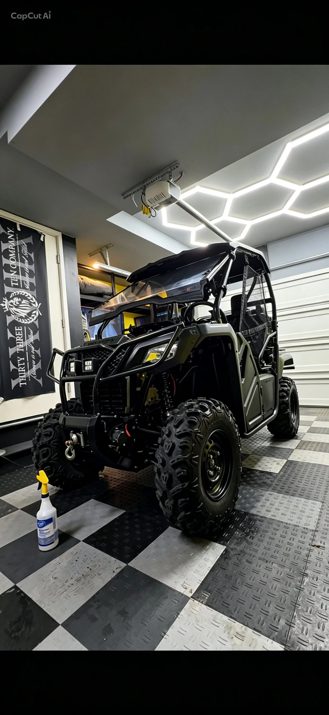 A black off-road utility vehicle parked in a garage with checkered flooring, a spray bottle on the floor, a hanging banner on the wall, and LED hexagon ceiling lights.