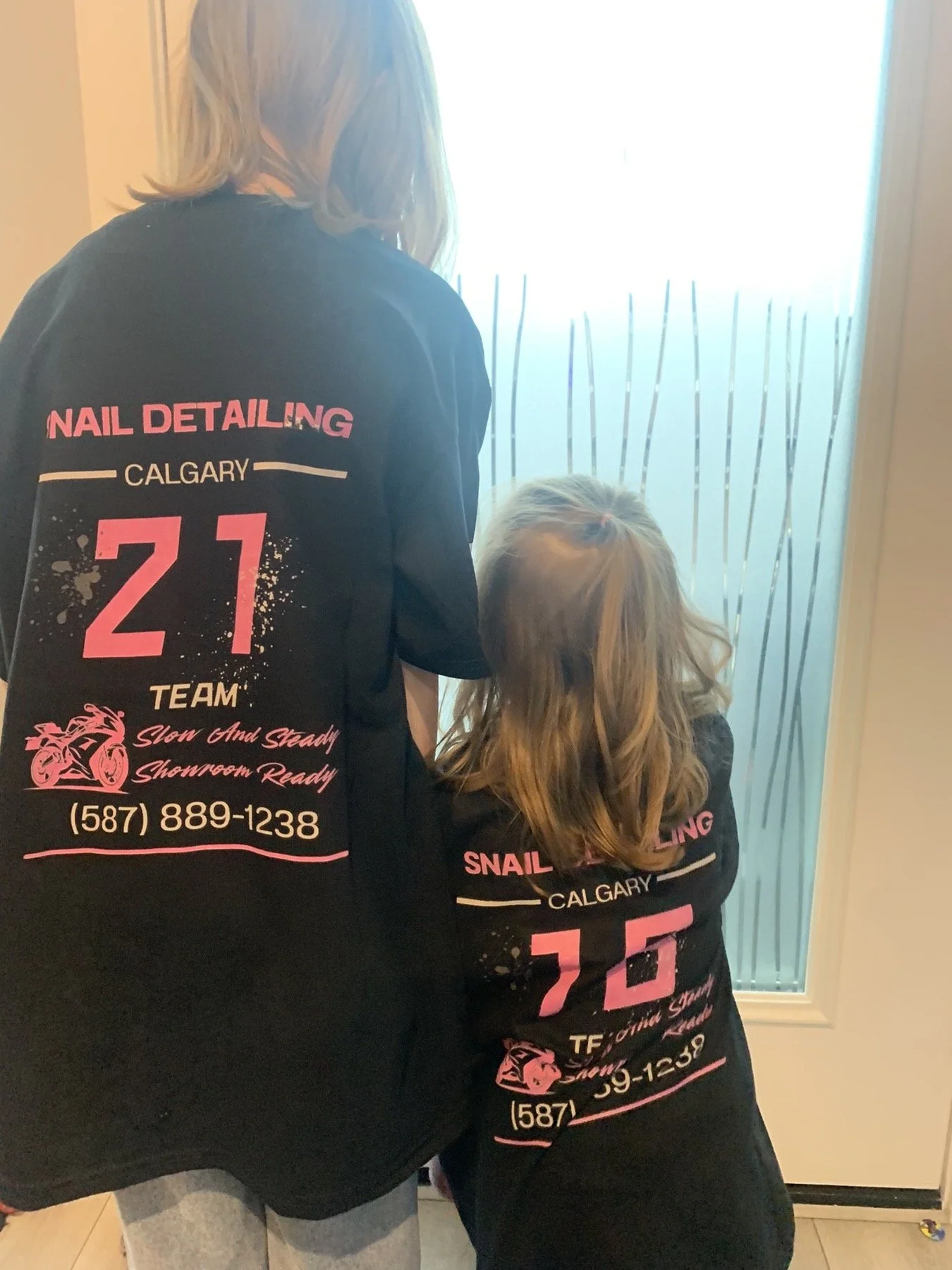 Two women, one taller and one shorter, wearing matching black T-shirts with pink and white text, standing indoors near a frosted glass door. The shirts promote nail detailing services in Calgary, with a phone number and graphic of a motorcycle on the back. The taller woman has light brown or blonde hair, and the shorter woman has long, dark hair.