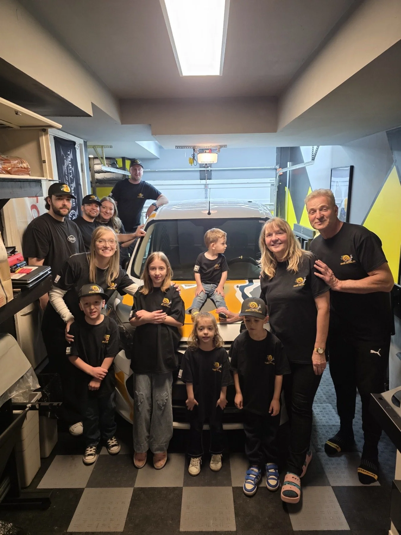 A group of children and adults gathered in a garage, posing around a yellow and white sports car with a tiger stripe design. The children and adults are wearing black shirts with a yellow logos, and some are wearing caps. There is a garage door and shelves with tools and equipment in the background.