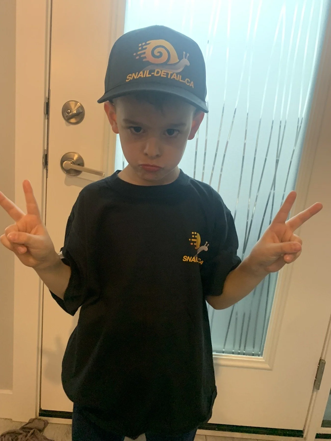 A young boy wearing a black T-shirt and a cap with the logo 'Snail-Detail.ca' is making peace signs with both hands in front of a door with frosted glass.