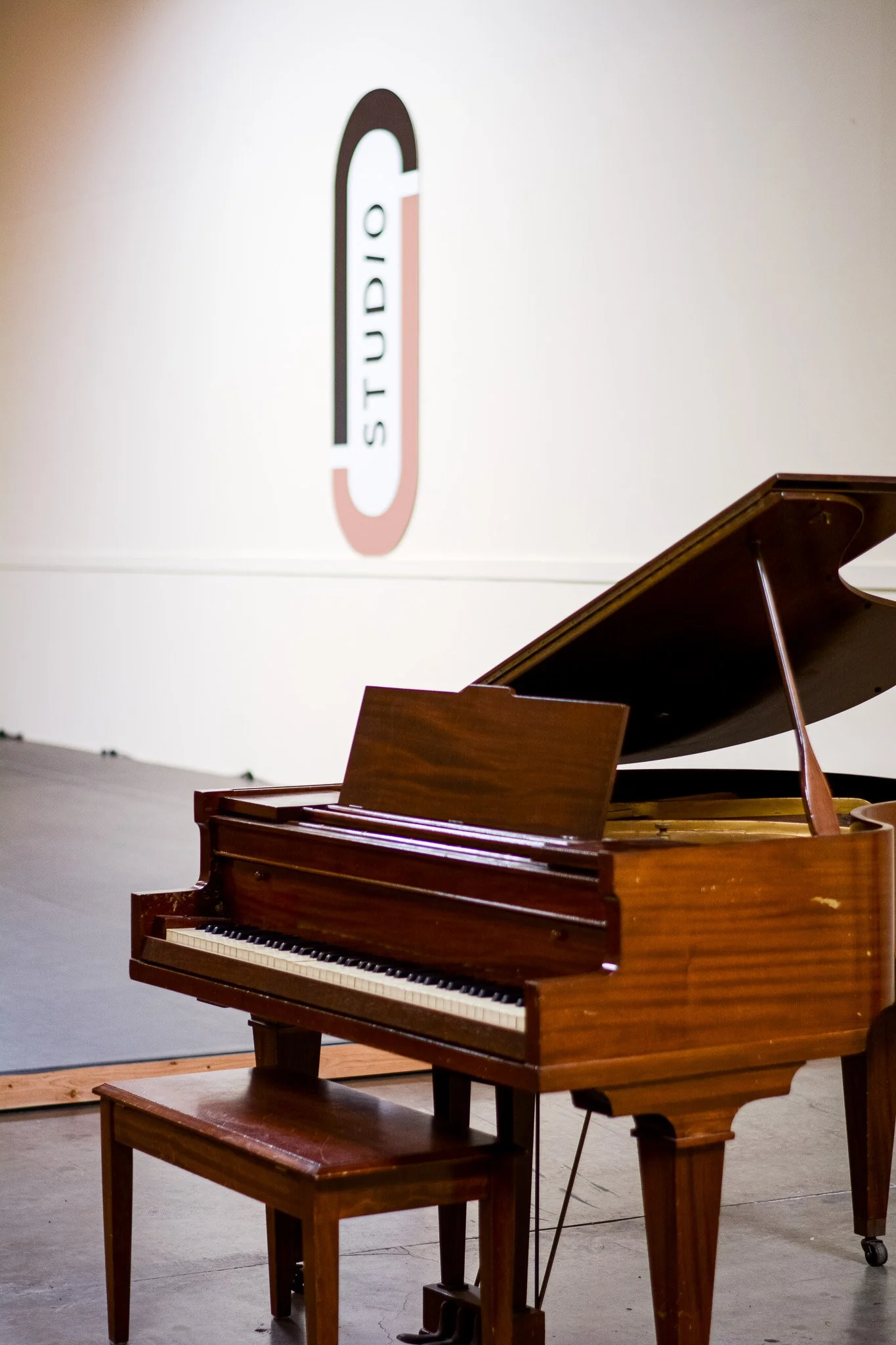 A wooden grand piano with a matching bench in a studio with white walls- studio rentals at Studio J Dance collective in Brea
