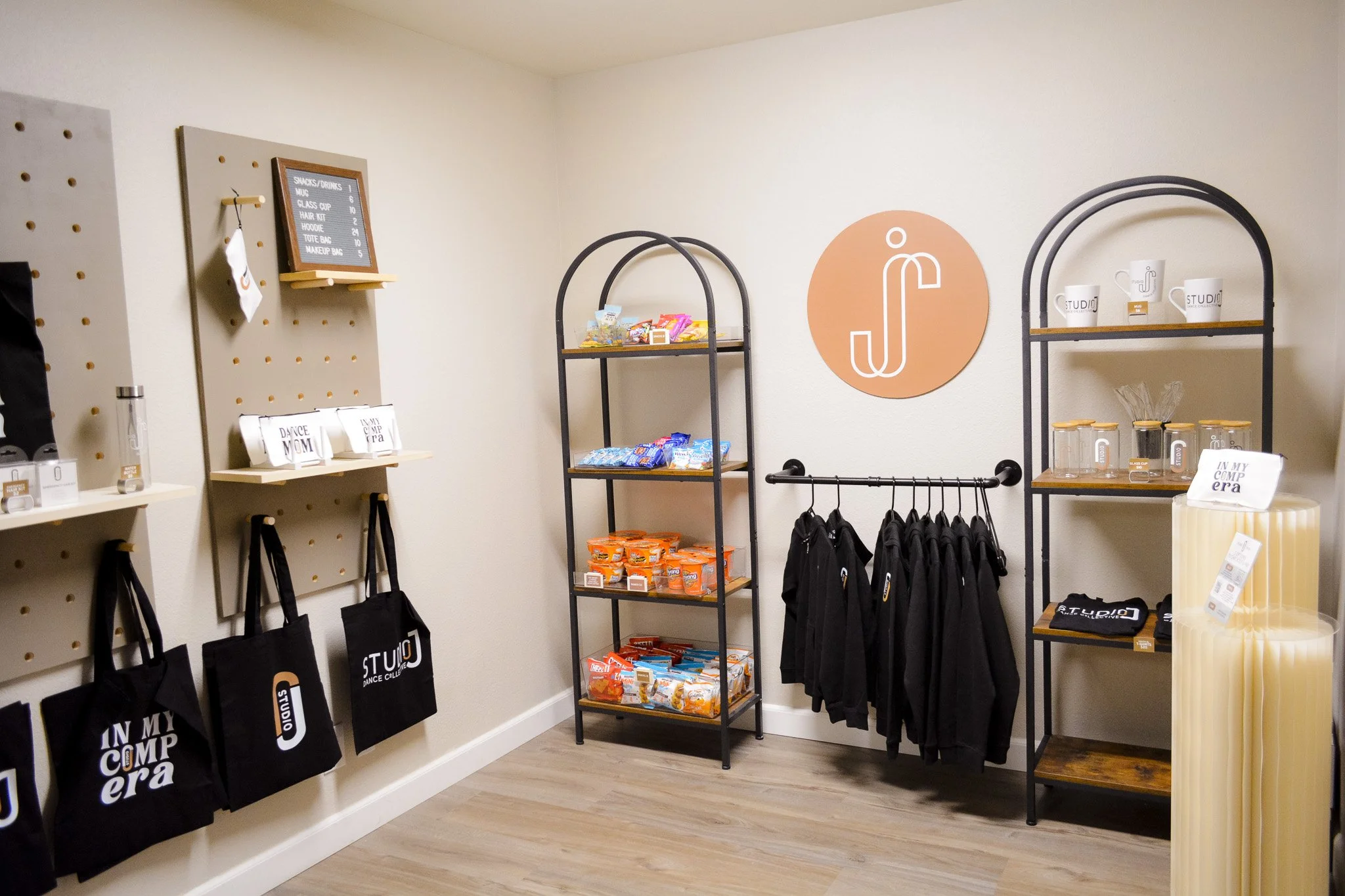 Store space with shelves of snacks, branded tote bags, dance merchandise with a dance studio logo, beige walls, and wooden flooring.
