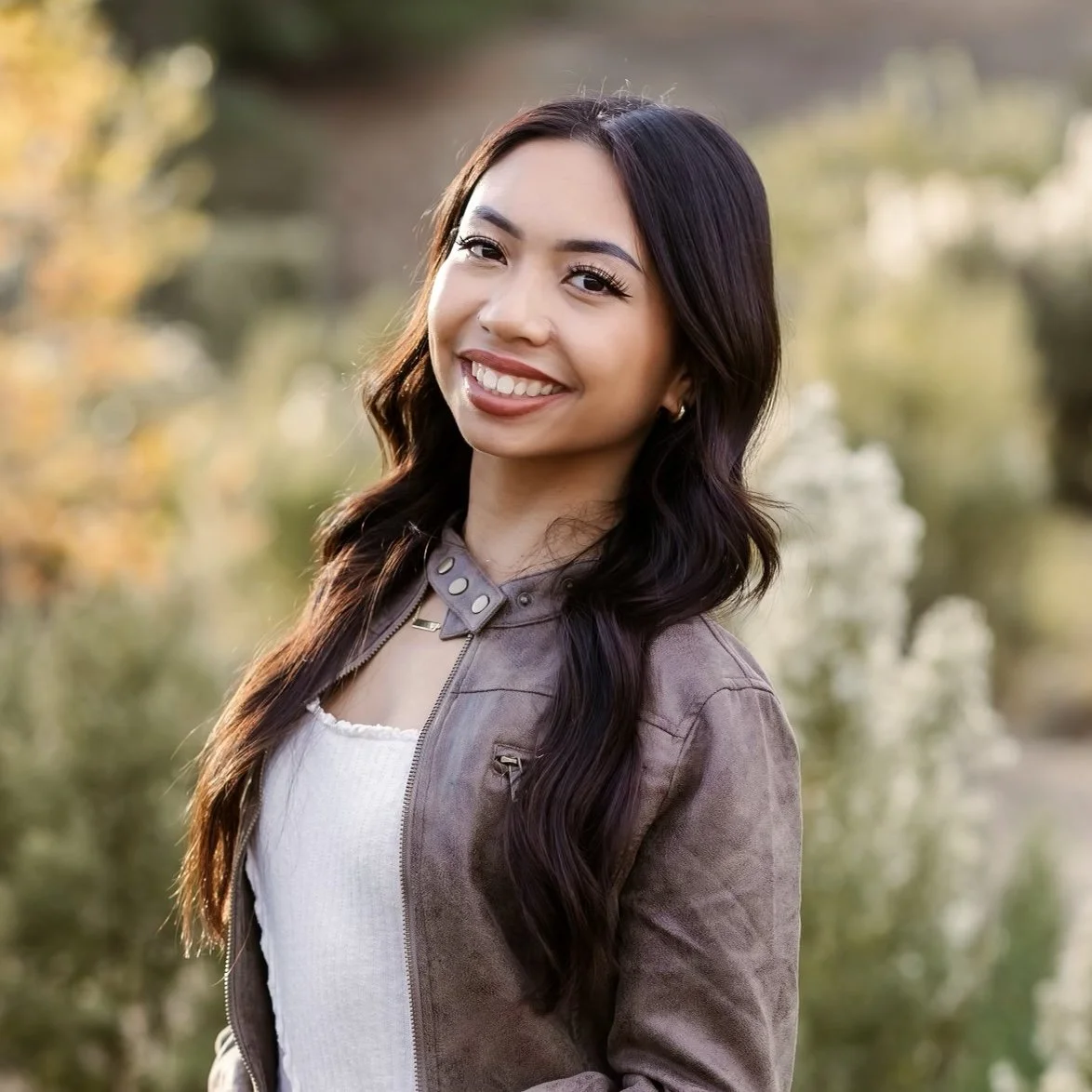 Young woman with long dark wavy hair smiling outdoors in a natural setting during daytime.